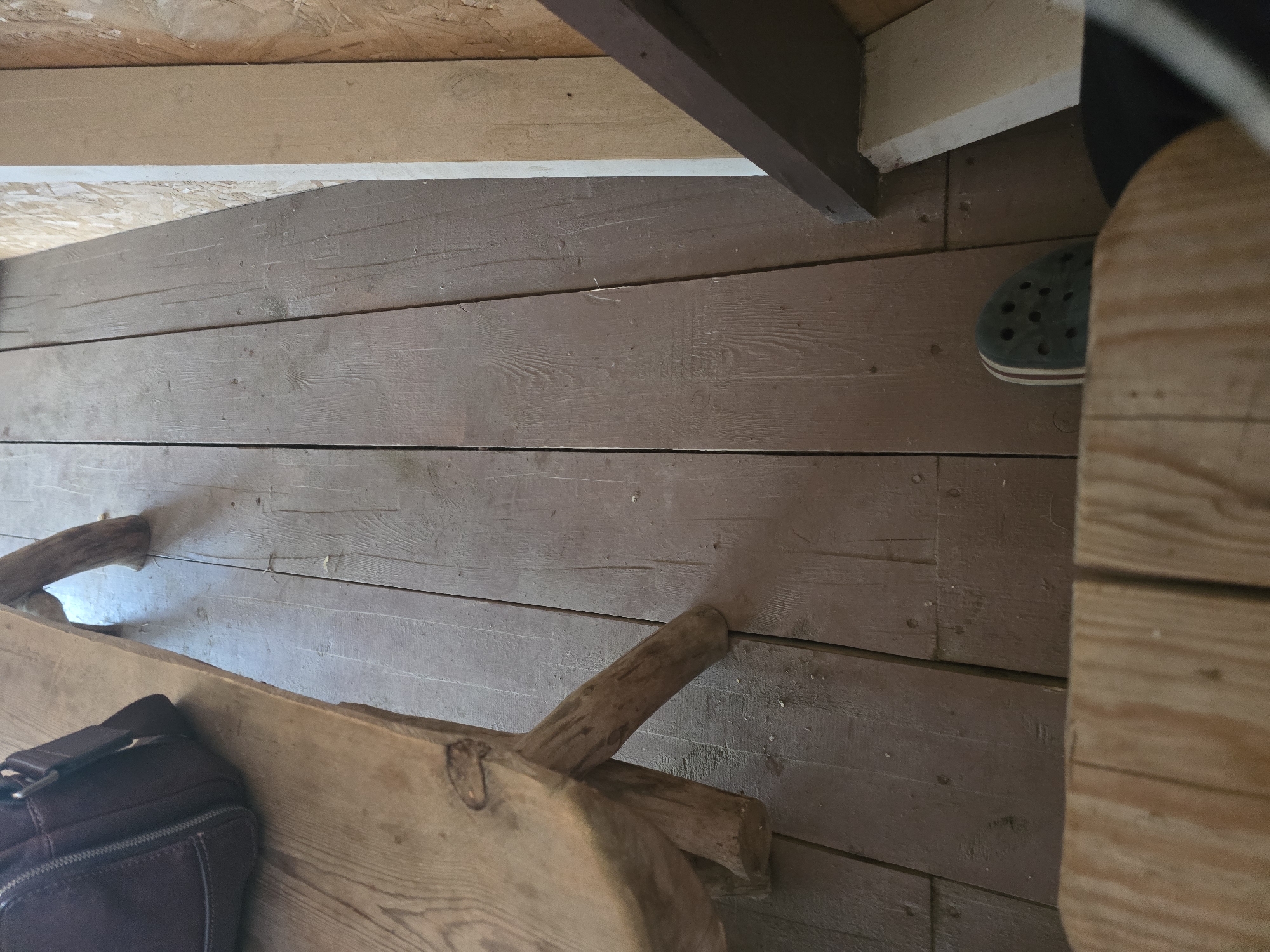 The image shows a close-up, low-angle view of a wooden floor with several wooden planks. The planks are oriented vertically from the viewer's perspective and have a worn, brown finish. There are visible nail heads and grain patterns in the wood.
On the left side of the image, a portion of a rustic wooden bench is visible, with a dark brown bag resting on its surface. The bench appears to be made of rough-hewn wood.
In the lower right quadrant of the image, a person's foot is visible, wearing a dark-colored clog-style shoe with red and white stripes on the sole.
The background on the right consists of a wall constructed of oriented strand board (OSB) and a vertical wooden beam. A dark, angled wooden structure is also present in the lower right, obscuring part of the wall and floor.
The scene appears to be an interior space with a rustic aesthetic. No specific location in Maksimiha, Russia can be determined from the visual information provided.