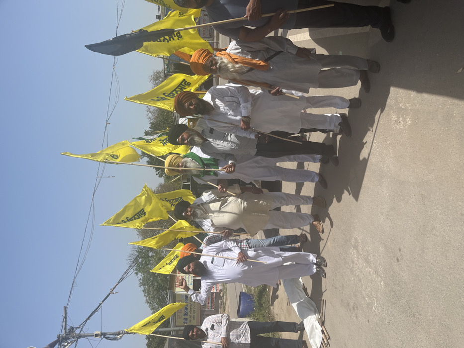 A group of 11 men, all wearing turbans, are standing in a line on a paved road, holding yellow flags with black text that reads "Punjab." They are holding the flags in their right hands and are standing in front of a white sheet on the ground. The men are facing the camera and appear to be participating in a demonstration or a protest. The coordinates, 30.706 latitude and 76.736 longitude, place the scene in Sahibzada Ajit Singh Nagar, India. A black flag with white writing on it is in the background, as is a gray car. The environment is urban, with wires in the sky and buildings in the distance. The road is paved, and the sky is a light blue. The men are all wearing white shirts and pants, and they are standing in a line that is not perfectly straight. The scene is likely a political rally or protest. The men are standing in a line, and their arms are at their sides, The men are all of similar age. There is a sense of solidarity and purpose in the scene.