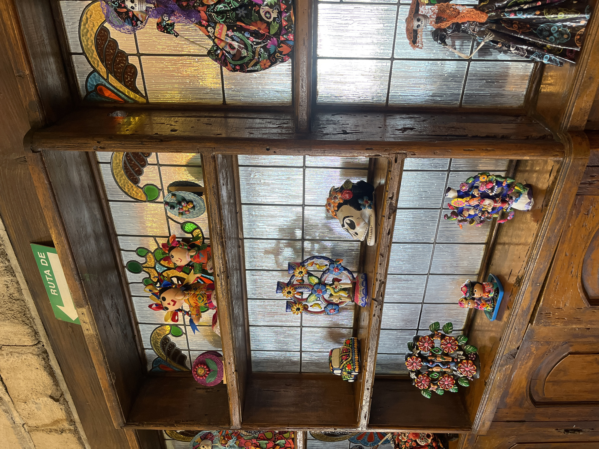 The image shows a low-angle shot of a decorative ceiling or shelf display in what appears to be a building in Puebla, Mexico. The ceiling is segmented into square or rectangular sections, each containing different ornamental items.
Several of the sections have decorative glass panes, some with colorful stained glass patterns. Among the items displayed are traditional Mexican folk art figurines, including dolls, ceramic skulls decorated with flowers, and other colorful ornaments.
A sign on the left side reads "RUTA DE" followed by an arrow, suggesting it's part of a directional sign for a tourist route or path. The overall environment evokes a sense of Mexican cultural decor.