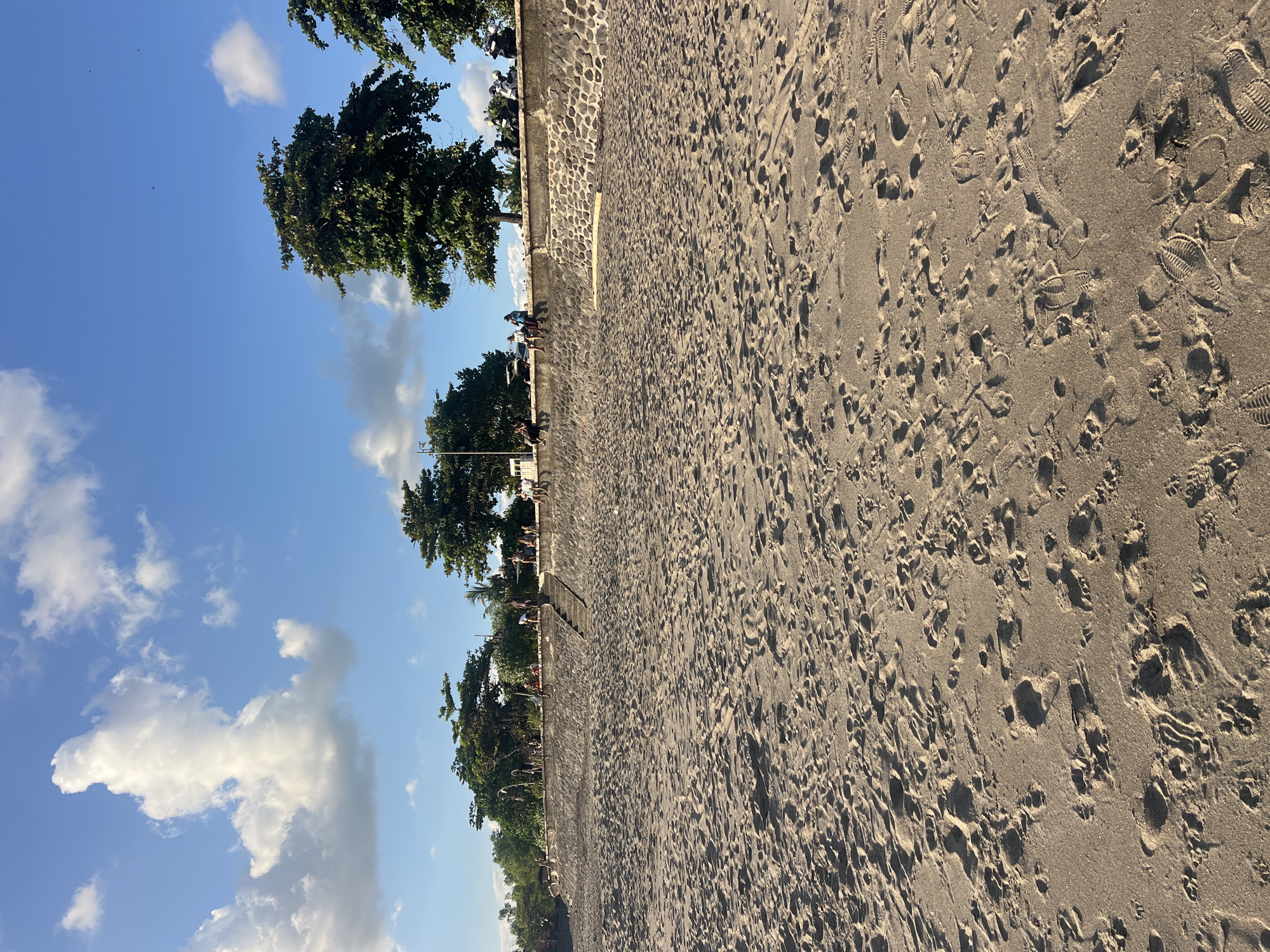 This image depicts a sandy beach in Kediri, Indonesia, with a stone wall bordering the sand. The sky is blue with scattered white clouds. Several people are visible sitting and standing on the wall, observing the scene. Lush green trees line the top of the wall, and beyond them, more vegetation is present. The sand is marked with numerous footprints, indicating recent activity. The overall impression is one of a pleasant day at a coastal location.