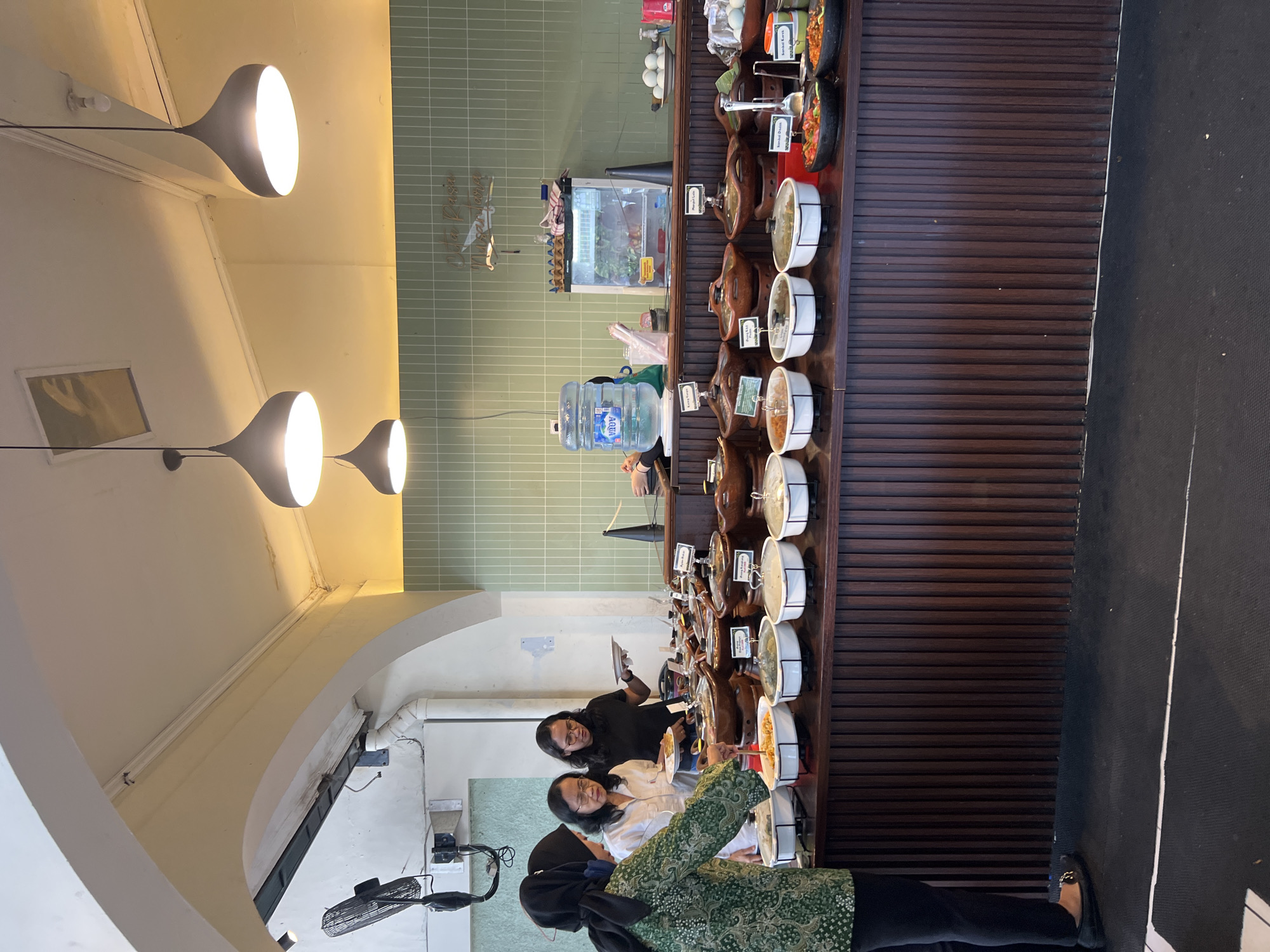 The image depicts a buffet-style food service area, likely in Yogyakarta, Indonesia, judging by the visible text on the wall sign which appears to read "Rasa Nusantara". The scene is set indoors, with a long wooden counter displaying numerous food dishes in clay pots and white bowls. The dishes appear to be a variety of Indonesian cuisine, including curries, stir-fries, and sambals.
In the foreground, three women are visible. The woman on the left, wearing a dark hijab and a green patterned traditional garment, is reaching into a white bowl with a serving spoon. The middle woman, wearing glasses and a white shirt, is holding a plate and looking at the food. The woman on the right, also wearing glasses and a black shirt, is holding a plate.
Behind the counter, there are several items including a large blue water dispenser, a small refrigerator with food inside, and a wall decorated with a green tiled pattern and a decorative sign. In the background, there is a ceiling with hanging lights and an industrial fan mounted on the wall. The overall ambiance suggests a casual dining establishment.