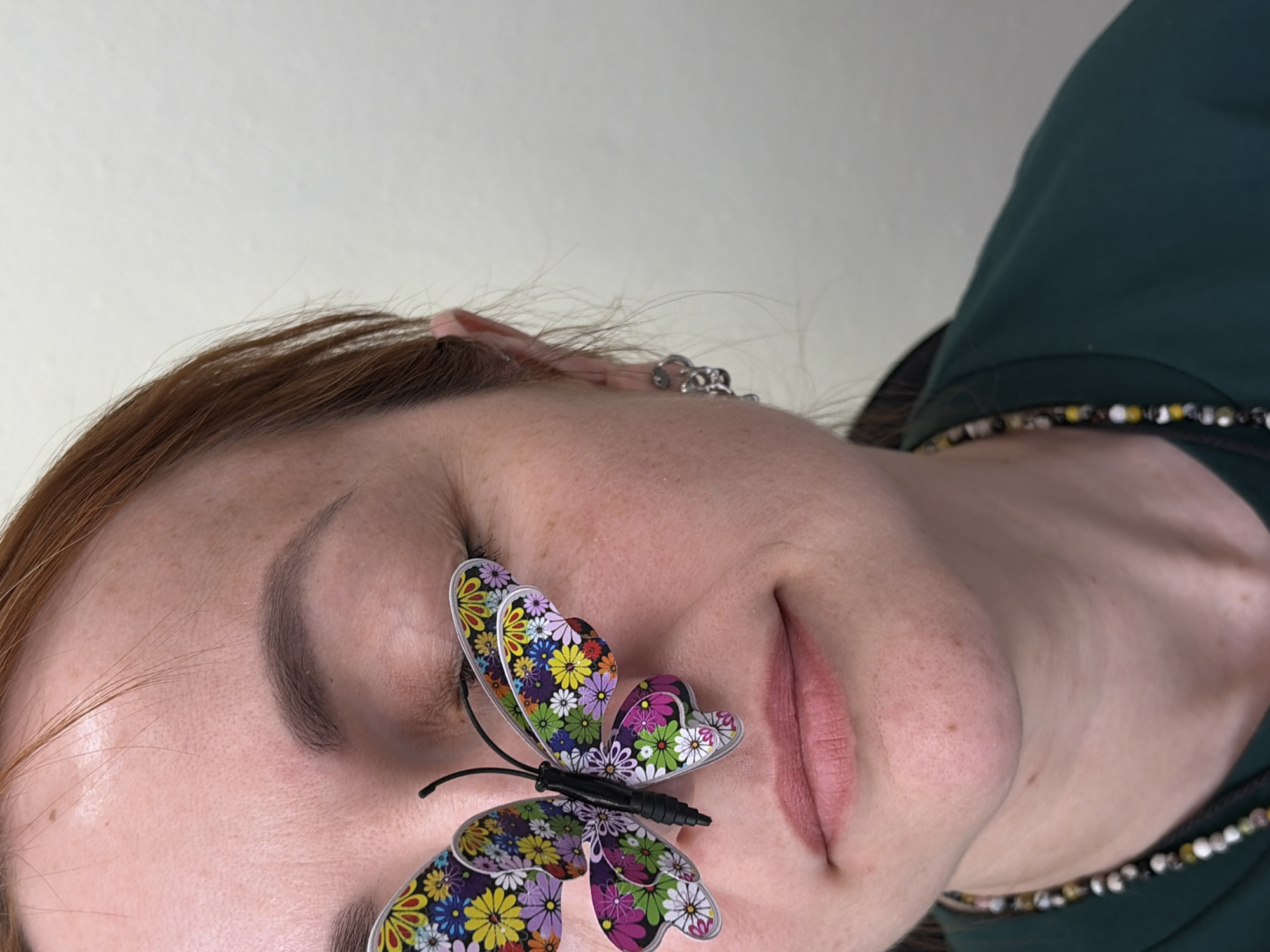 A medium close-up photograph shows a light-skinned individual with closed eyes, a butterfly sticker affixed to their forehead. The individual has reddish-brown hair, a defined eyebrow, and light pink lips. An earring can be seen on the ear. The butterfly sticker is multi-colored with a floral pattern. The person wears a dark green garment with a beaded necklace. The background is a plain, light-colored wall. The city of Chaikovsky, Russia, cannot be determined from this image.