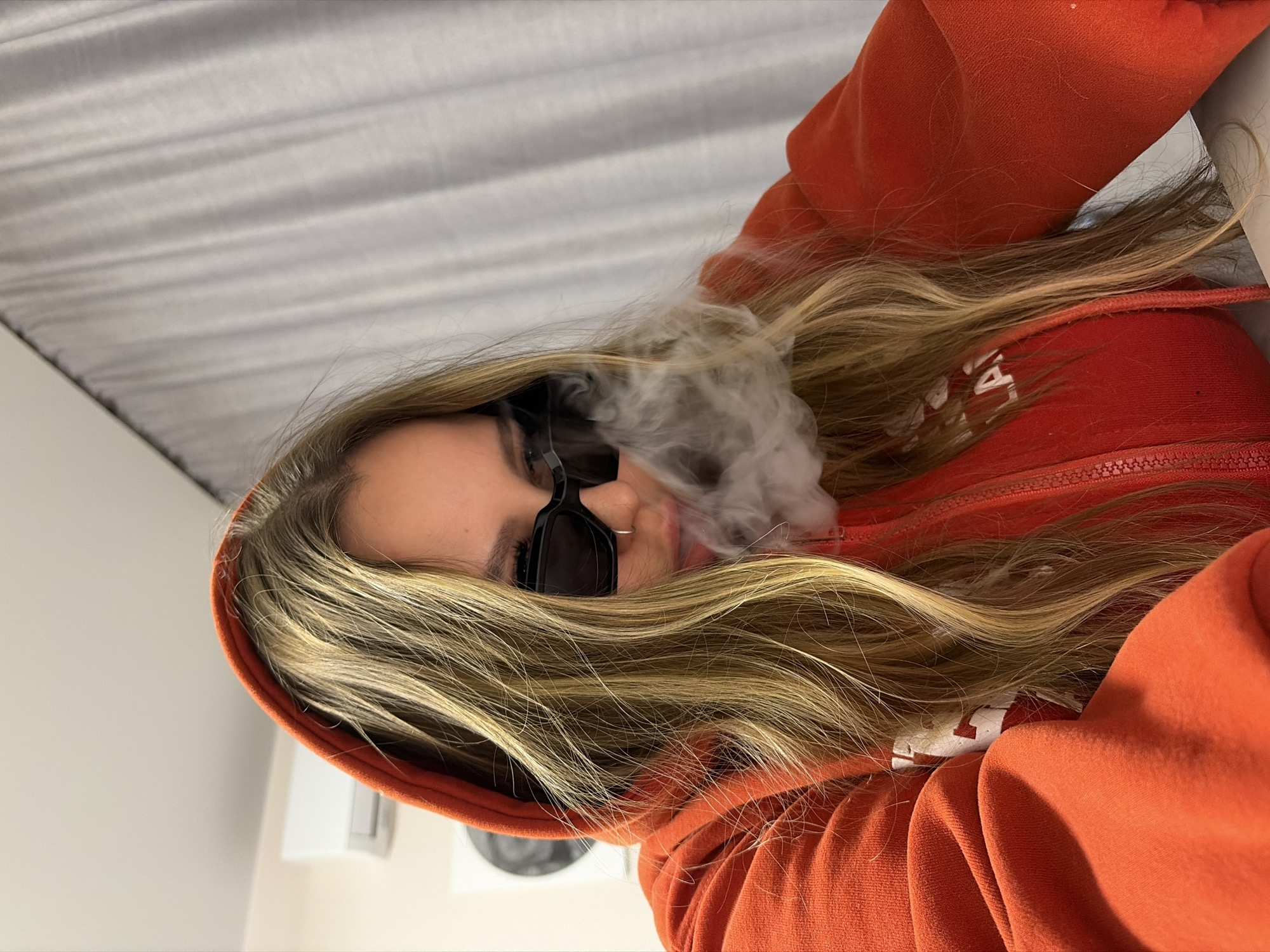 A young woman with long blonde hair and sunglasses is wearing an orange hooded sweatshirt. She is partially exhaling a cloud of white vapor, possibly from smoking or vaping. The background is blurry, showing a white ceiling or wall. The location is not discernable from the image, therefore the city of Krasnodar, Russia is unknown.