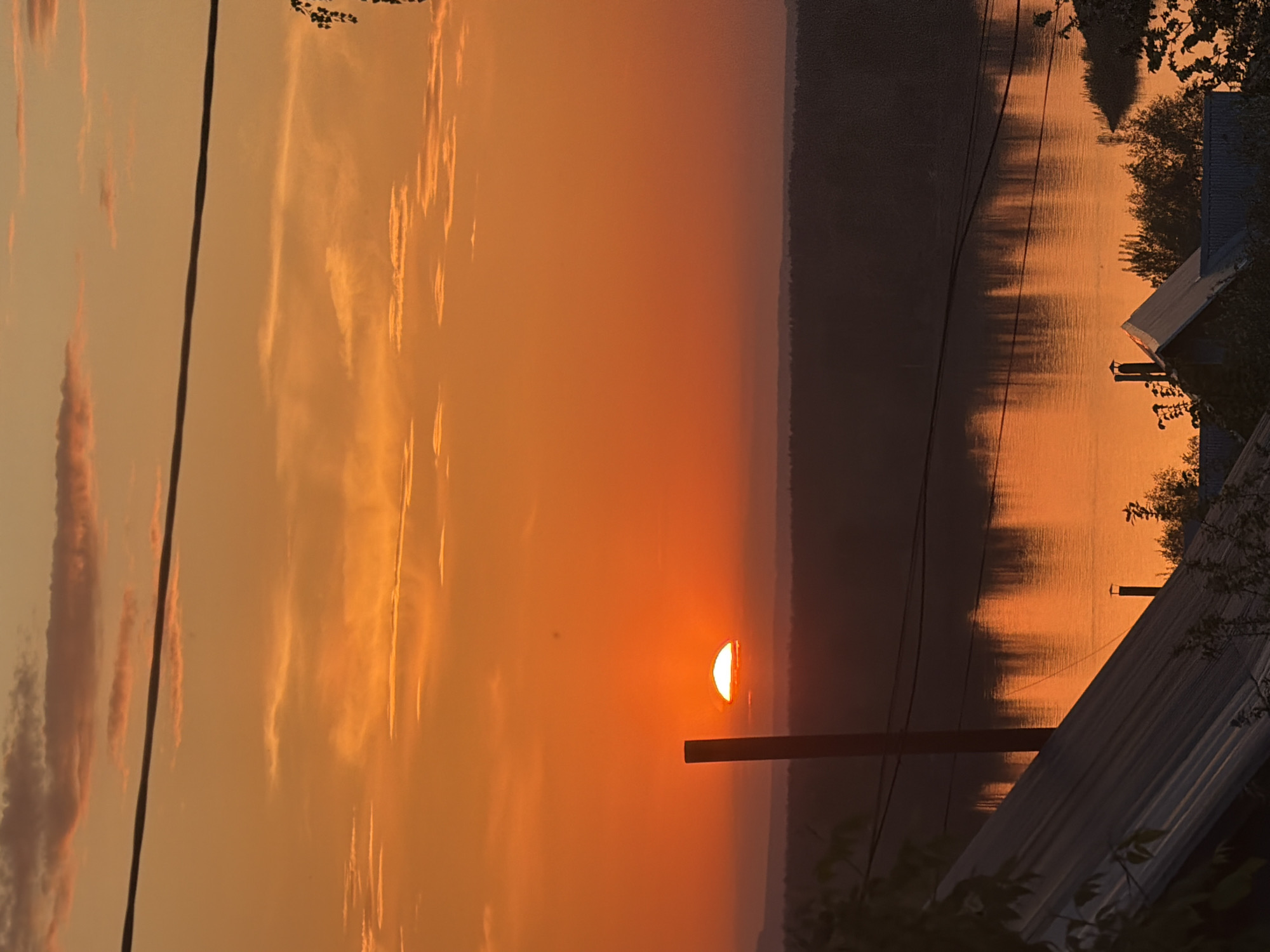 The image depicts a sunset over a body of water, likely a river or lake, with the sun partially obscured by the horizon. The sky exhibits orange and yellow hues, indicating either sunrise or sunset. Reflections of the sky and the horizon are visible on the water's surface. Several utility poles and power lines are present in the foreground. Parts of a building with a metallic roof are on the edge of the image. Trees or bushes are on the right edge of the image. The location is likely Chaikovsky, Russia, based on the information provided.