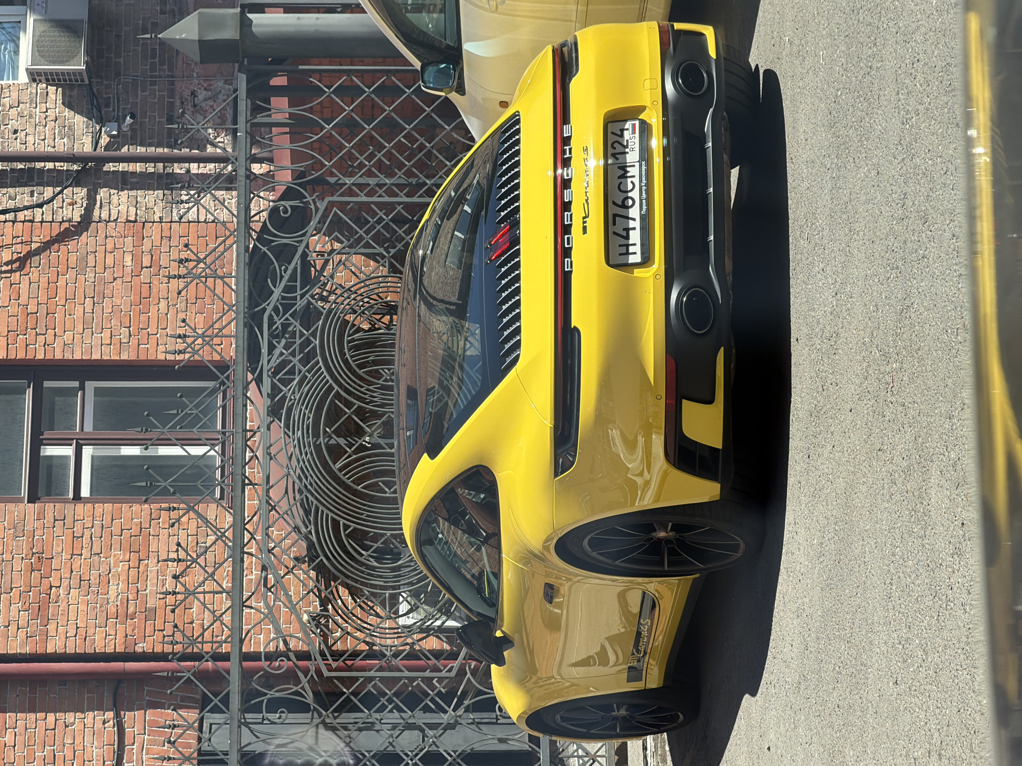 A bright yellow Porsche 911 Carrera S is parked on a paved surface in front of a decorative metal gate and a red brick building. The car is viewed from its rear-right quarter. Its license plate reads "H476CM 124 RUS" with the text "Порше Центр Красноярск" below it, indicating it is located in Krasnoyarsk, Russia. The car has a black roof and black wheels. The building behind the car features a window and a red pipe running down its facade. Sunlight casts shadows on the ground.