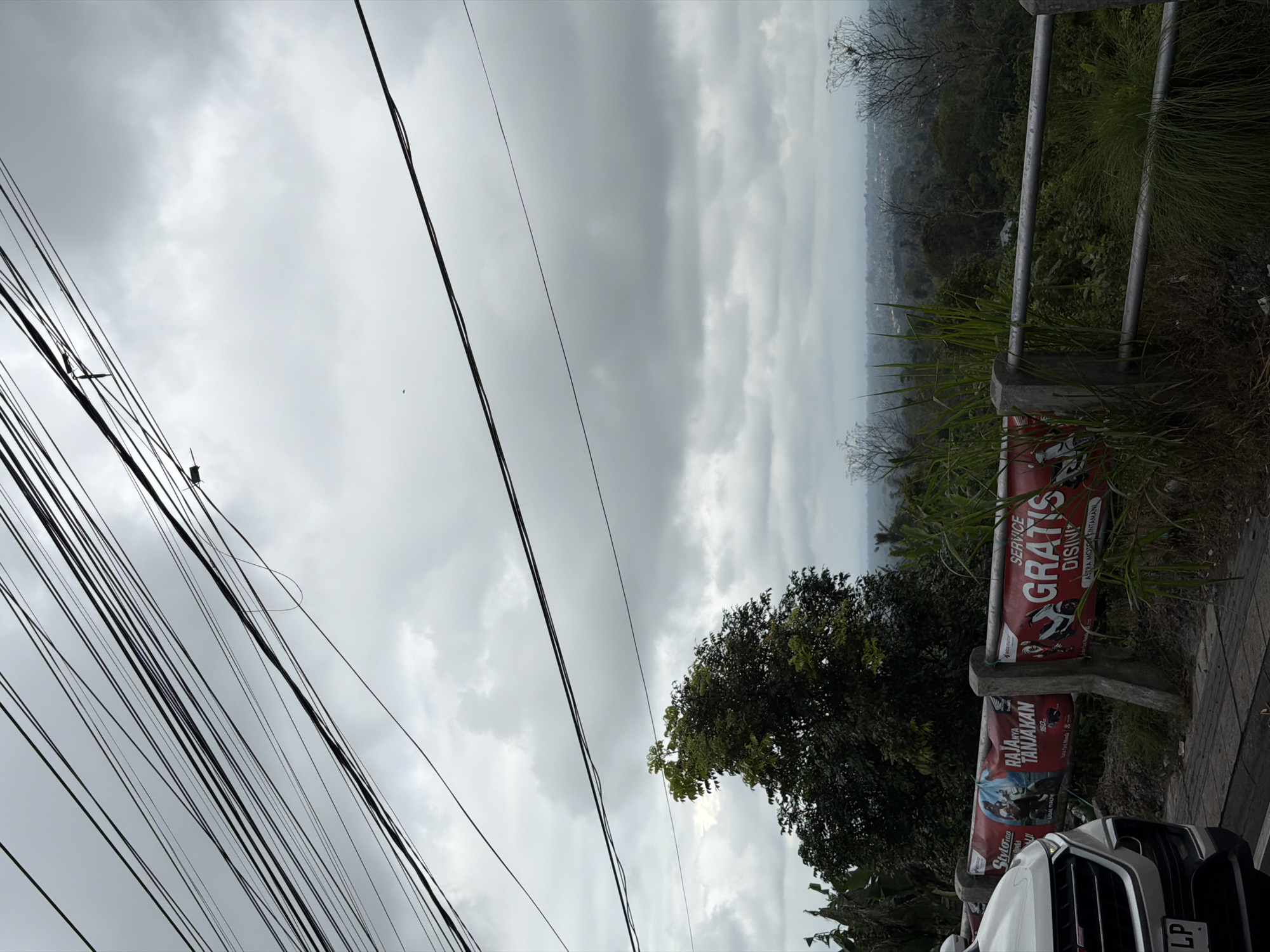 This is a photograph taken in Kintamani, Indonesia, on a cloudy day. The foreground shows the front of a white car with a license plate that reads "LP". To the right of the car, there is a concrete barrier with a metal railing, overgrown with grass and foliage. Several red banners are attached to the barrier. One banner has an image of a motorcycle and text that appears to say "RAJA-NYA TANJAKAN". Another banner displays the text "SERVICE GRATIS DISINI". Behind the barrier, a lush green landscape with trees and distant buildings is visible under a heavily overcast sky. A dense canopy of power lines stretches across the upper portion of the image, obscuring the sky.