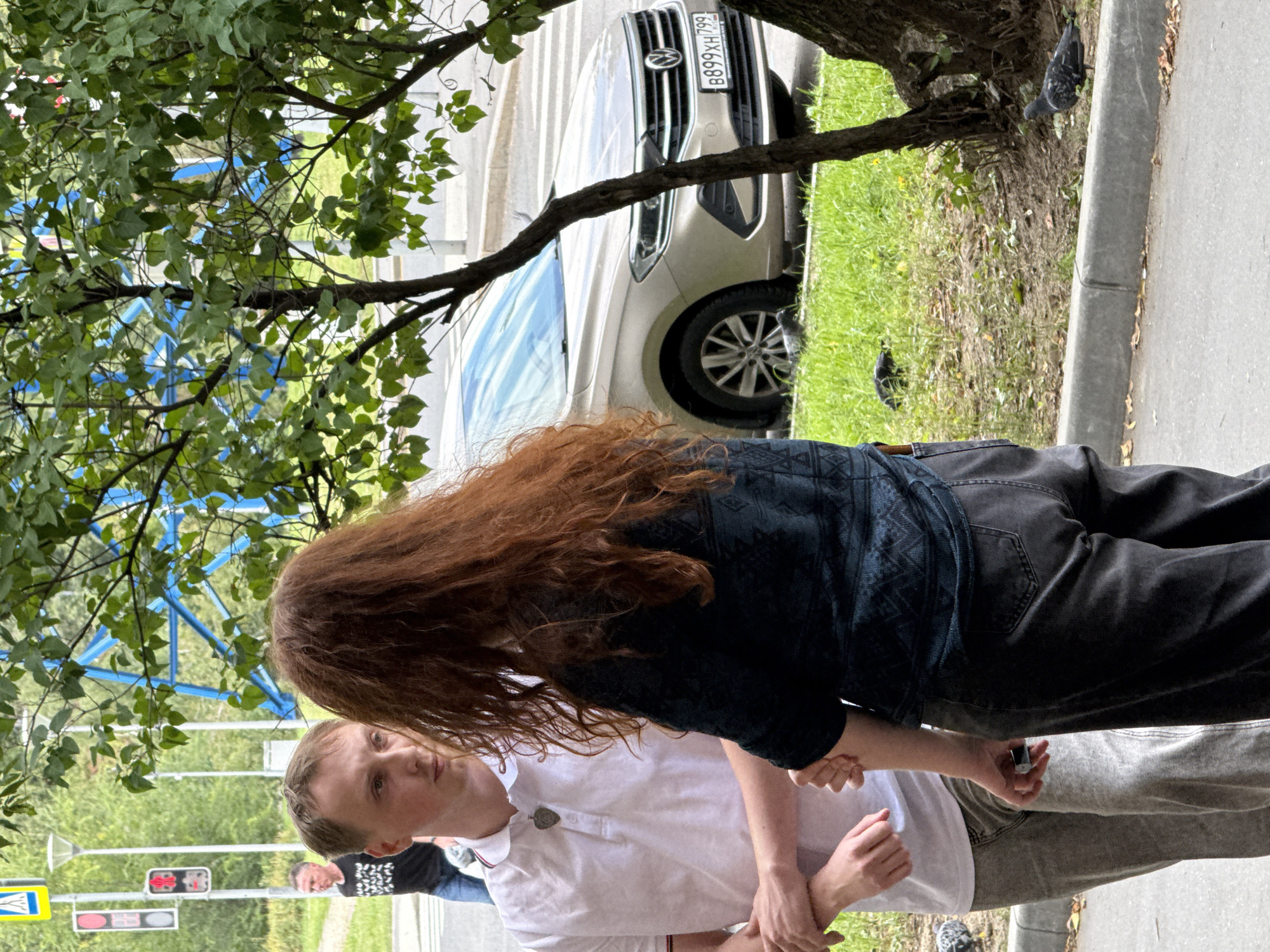 Two individuals, a man and a woman with long red hair, stand facing each other in close proximity on a sidewalk in Moscow, Russia. The man, on the left, wears a white polo shirt and grey trousers. The woman, on the right, is dressed in a dark patterned top and dark trousers. Their arms are intertwined, suggesting an intimate interaction. Behind them, a light-colored Volkswagen sedan is parked on the street. The environment includes green grass, trees, and traffic signals in the background, indicating an urban setting.
