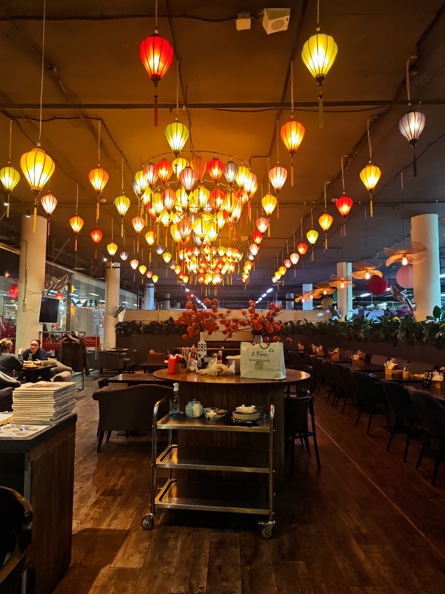 This image depicts the warm and inviting interior of a restaurant or cafe in Moscow, Russia. The dominant feature is the ceiling adorned with numerous traditional Asian-style lanterns in various shades of red, orange, and yellow, creating a vibrant and festive ambiance. These lanterns are suspended at different heights, forming a large, central cluster and extending in rows along the length of the space.
The restaurant features dark wooden floors and a mix of dark wooden tables, chairs, and upholstered seating. On the left side of the foreground, a stack of magazines or newspapers rests on a counter. Two individuals are seated at a table nearby, one facing away from the camera and the other with dark hair, possibly engaged in conversation or looking down. In the mid-ground, a wooden table holds a vase with red decorative branches or berries and a white paper bag. The text "A Thanh" (partially visible) and "RANOCCHIO MAISON COFFEE" can be read on the paper bag. A metal serving cart with two shelves stands in the center foreground.
Further into the background, more tables and chairs are arranged. On one table, a small orange sign with the number "03" is visible. The ceiling is modern and somewhat industrial, with large white pillars. Beyond the seating area, glimpses of an adjacent indoor space, possibly a shopping mall or large complex, are visible through glass partitions. The lighting is dim, suggesting it is evening or night, relying entirely on the glow from the lanterns and other internal fixtures. The overall scene conveys a relaxed and distinctly themed dining environment.
