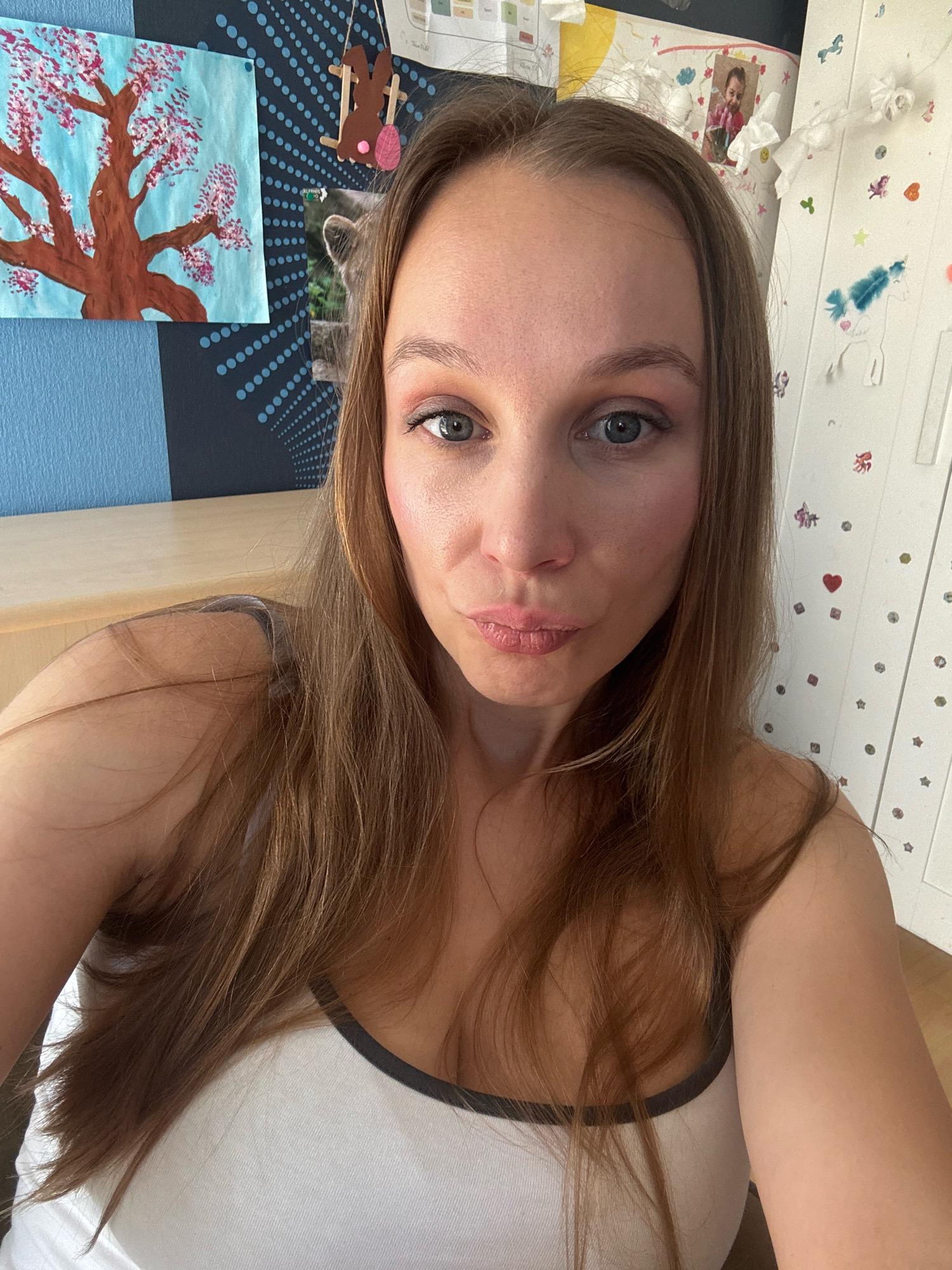 A woman takes a selfie in a room. She has long, brown hair and is wearing a white tank top. Her makeup includes eyeshadow and lipstick. In the background, there is a drawing of a tree with pink blossoms, a piece of blue fabric, and a white cabinet decorated with stickers of stars and unicorns. The room appears to be a child's bedroom or a creative space. The overall atmosphere is casual and personal. The image does not contain enough information to determine the specific location in Überherrn, Germany.