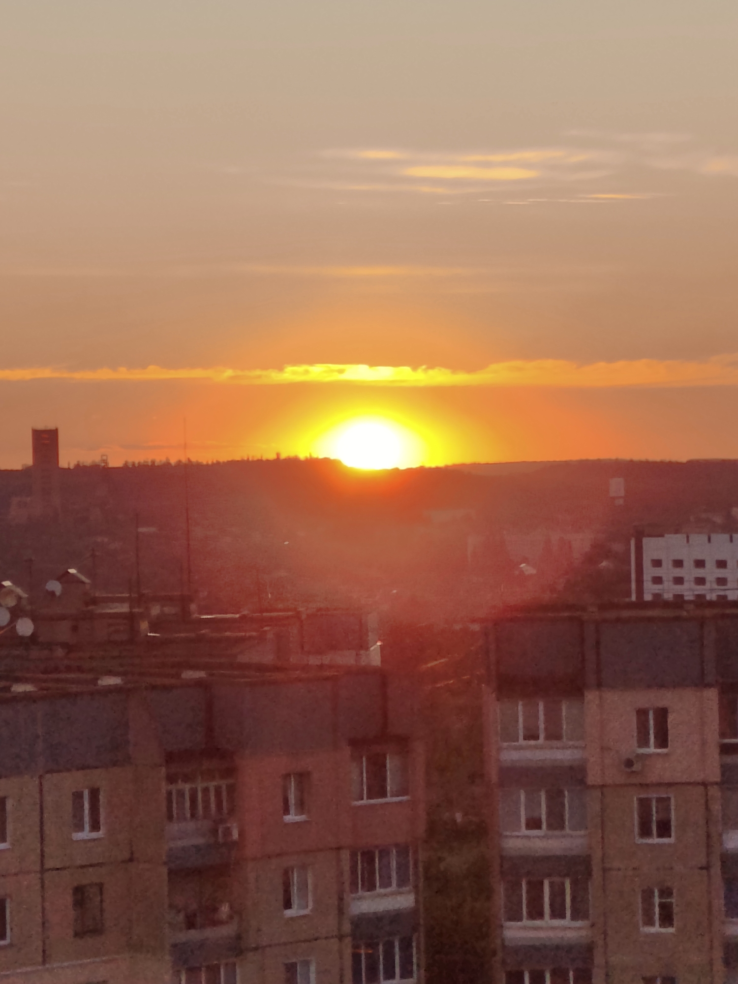 The image depicts a sunset over the city of Kryvyi Rih, Ukraine. The sun, a bright orange orb, is low on the horizon, casting a warm glow across the sky and the landscape below. Wispy clouds are scattered across the sky, tinged with orange and yellow hues. In the foreground, the rooftops of apartment buildings are visible, some with satellite dishes attached. The buildings appear to be made of brick and concrete, with a muted color palette. In the background, a tall, slender tower stands out against the skyline, its silhouette dark against the colorful sky. The overall scene is one of a tranquil evening in an urban environment.