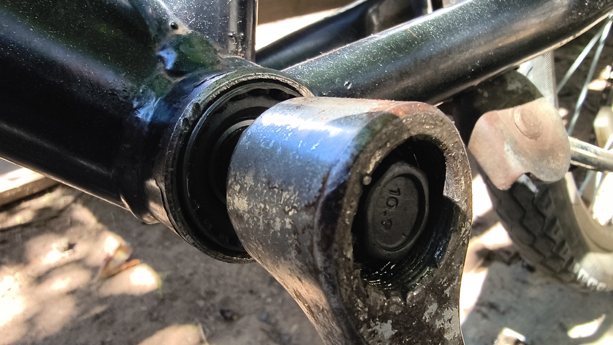 The image shows a close-up, low-angle view of a bicycle's crank arm and bottom bracket. The crank arm, made of metal and showing signs of wear and dirt, is attached to a black metal frame. A bolt with "10.9" stamped on it secures the crank arm. Visible within the frame is a bearing assembly, also dirty. To the right, a portion of the bicycle's rear wheel, including the tire and spokes, is visible. The background is out of focus, showing what appears to be a ground surface with some dappled sunlight. The scene is likely outdoors, possibly in a workshop or residential area. No people or specific identifying landmarks of Mikhaylovka, Russia, are present in the image.