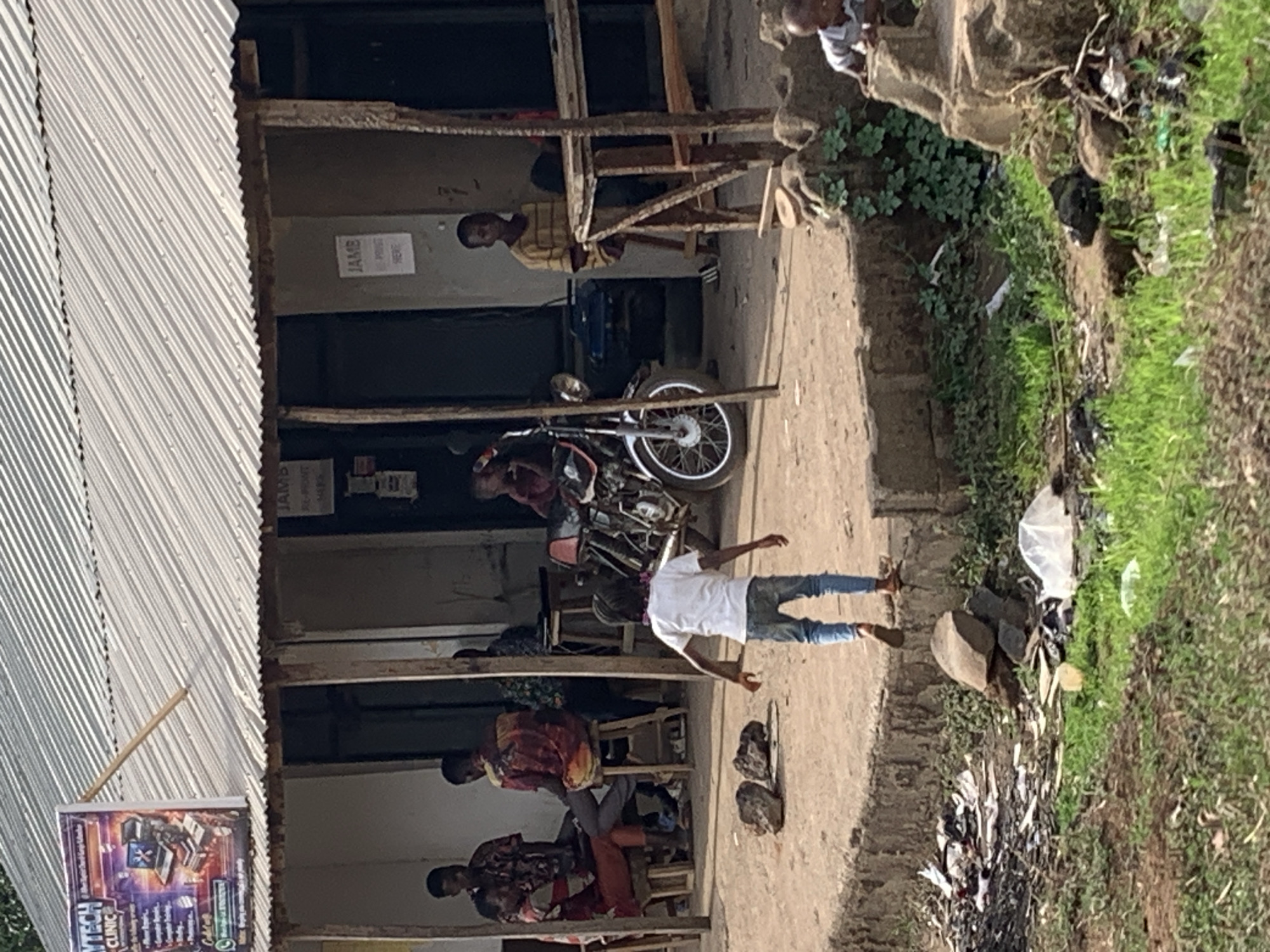 The image depicts an outdoor scene in Agban, Nigeria. Several people are sitting under a corrugated metal roof structure supported by wooden beams. A motorcycle is parked nearby. One individual stands outside the structure. A small sign labeled "JAMB" is visible on the wall. A second sign with the text "MYTECH CLINIC" is posted on the side of the structure. The ground is bare earth, and trash is scattered along the edge of the area bordering green vegetation.