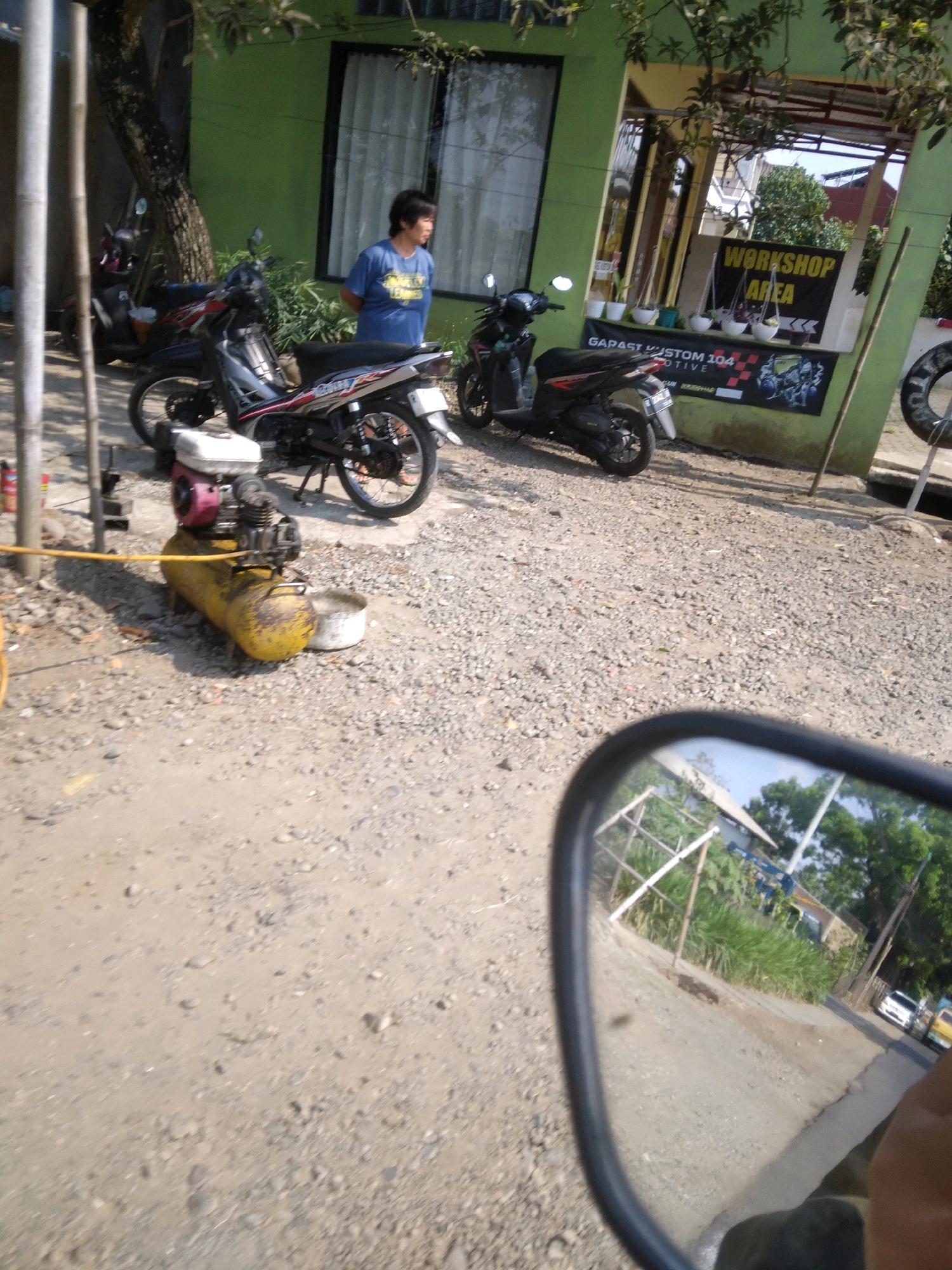 In Desa Pamijen, Kecamatan Sokaraja, Indonesia, a scene unfolds in front of a green building with a covered entrance. A person wearing a blue t-shirt stands near the entrance, looking towards the right. Two motorcycles are parked in front of the building, one black and red, and another black scooter. A yellow air compressor with a black engine sits on the ground to the left of the motorcycles. The ground is covered in gravel and dirt. In the reflection of a motorcycle's side mirror, a road with passing cars and vegetation can be seen. The overall environment suggests an outdoor workshop or roadside business.