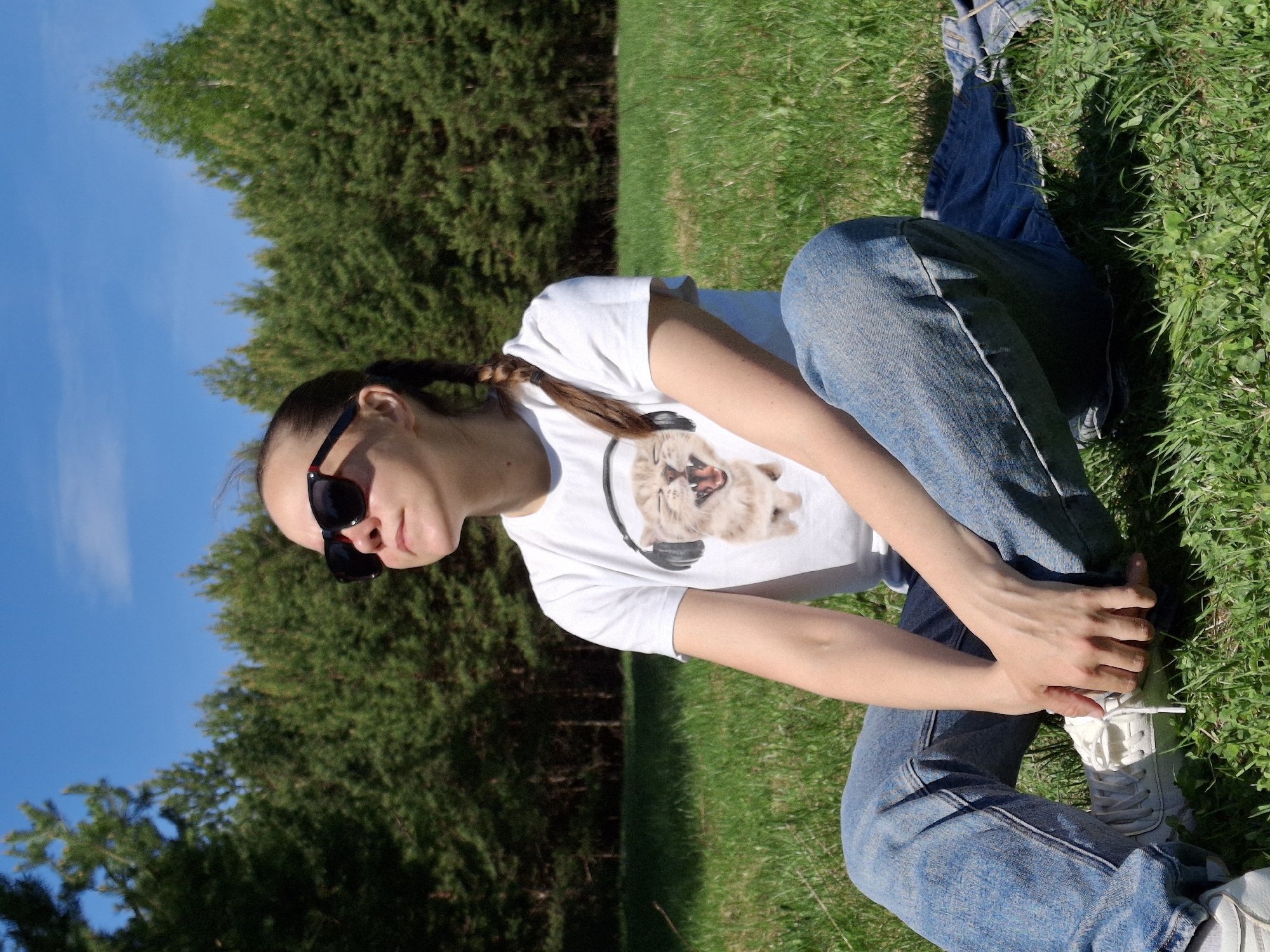 A woman with a brown ponytail, wearing sunglasses, a white t-shirt with a cat wearing headphones graphic, and blue jeans, is sitting cross-legged on a grassy area. She is holding the laces of her white sneakers. Behind her are green trees and a blue sky. Discarded denim fabric is near her on the grass. The location is likely Chaikovsky, Russia, as indicated by the prompt.