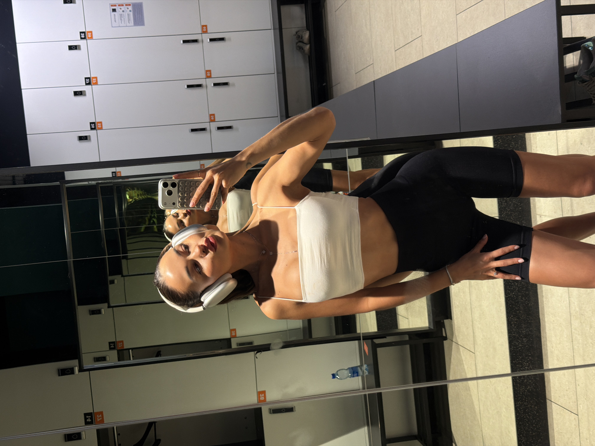 A young woman is captured in a mirror selfie within a gym changing room located in Moscow, Russia. She stands centered, facing her reflection while holding a light-colored smartphone with a distinct patterned case featuring circular indentations in her right hand. She is wearing white over-ear headphones, a white spaghetti-strap crop top, and black bike shorts. Her dark hair is pulled back, and she has a necklace with a drop pendant, a silver bracelet on her right wrist, and a ring on her right ring finger. Her lips are puckered in an expressive pose.
The background, visible through the mirror, reveals white lockers with black handles and small orange labels displaying reversed Arabic numerals (e.g., "39", "40", "38") and reversed Cyrillic text beneath them. Below the lockers, a dark-colored bench or shelf extends across the scene. On the left side of the reflection, a blue plastic water bottle rests on the bench. On the right, a pair of light-colored athletic shoes is visible on the floor beneath the bench. The floor consists of light-toned rectangular tiles with darker dividing lines. A vertical metal strip divides the mirror panels on the left side of the image. The upper portion of the background reflects dark surfaces, possibly the ceiling or upper walls of the changing room.
