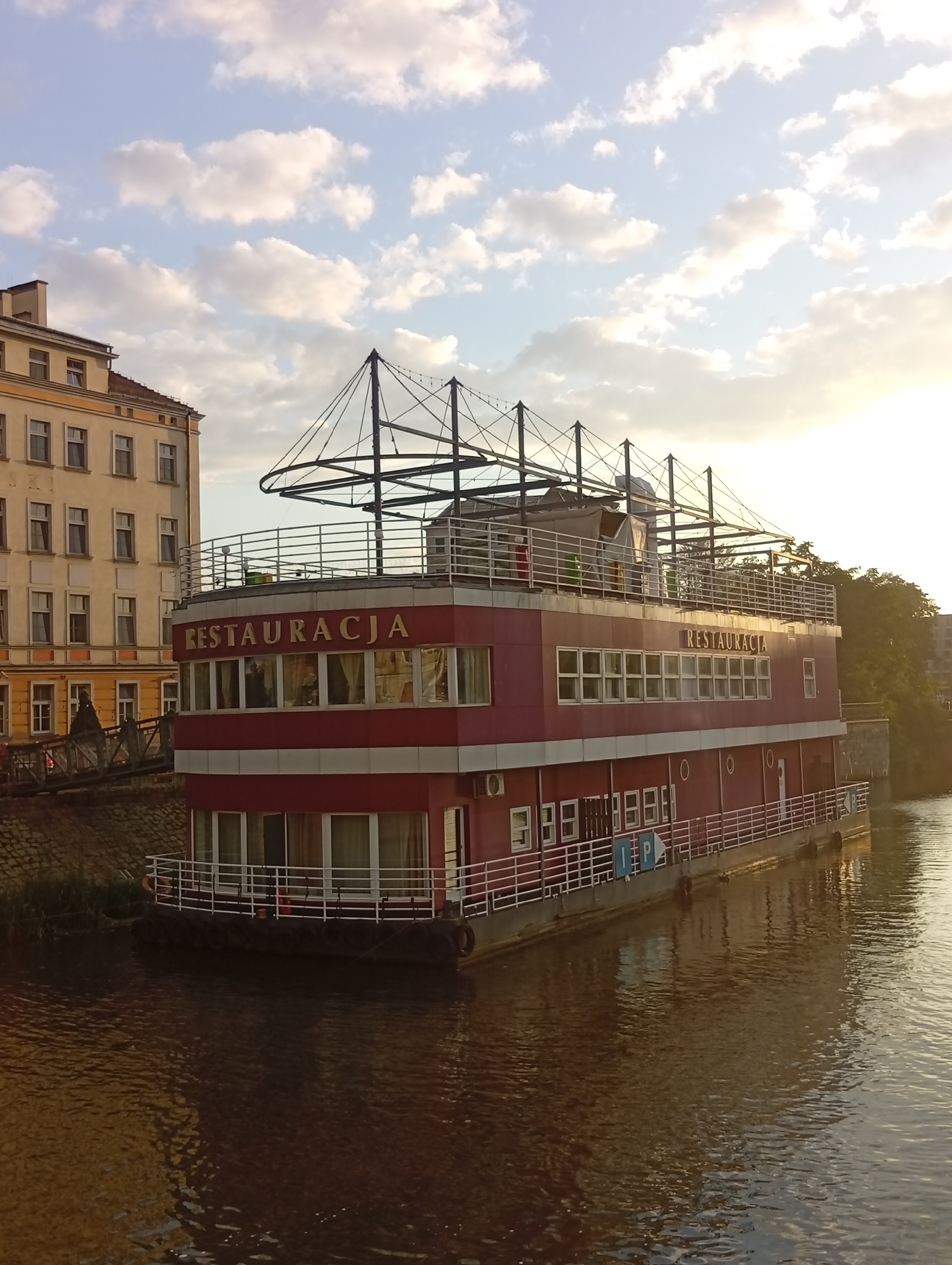This is an exterior, medium-shot, eye-level, still photograph of a large, red and white restaurant boat docked on a river in Wrocław, Poland. The boat, named "RESTAURACJA," is a multi-story structure with large windows on each level. The upper deck features an open area with a metal railing and a covered section. A ramp leads from the riverbank to the boat's entrance. The boat is surrounded by water, with reflections of the boat and the sky visible on the surface. To the left of the boat is a light-colored, multi-story building with numerous windows. The sky is filled with scattered white clouds against a pale blue background, with sunlight streaming in from the right side of the frame, indicating it is likely late afternoon or early evening. The environment appears to be an urban setting, given the presence of the building and the docked boat. There are no visible people or animals in the frame.