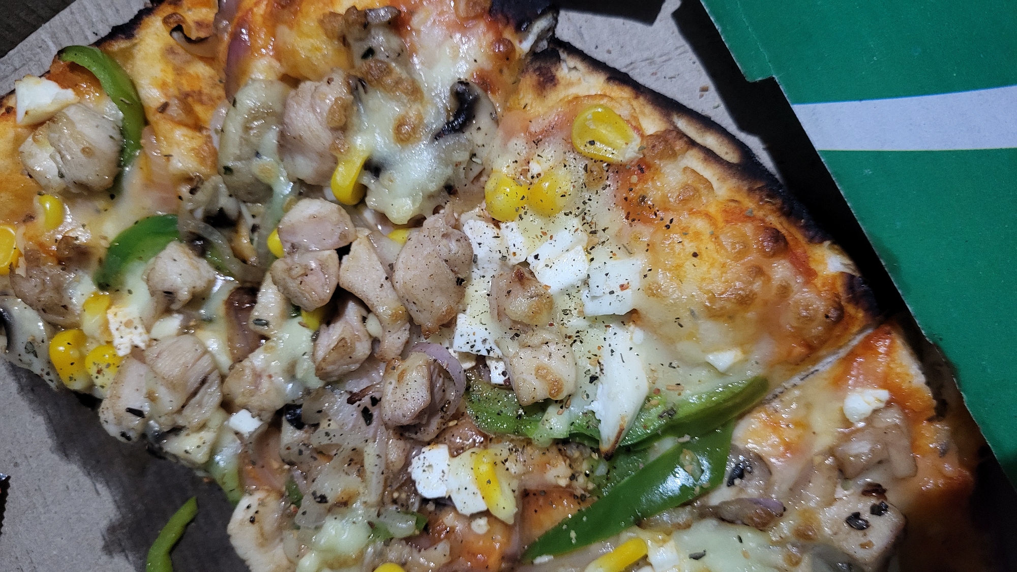 A close-up, high-angle view depicts a freshly baked pizza in a cardboard box, situated in the city of Vashisht, India. The pizza features a golden-brown crust with some charred edges. Toppings are generously distributed across the surface, including cubed pieces of cooked chicken, scattered yellow corn kernels, green bell pepper strips, visible slices of red onion, and white, diced cheese or paneer. The entire surface is covered with melted, bubbly cheese, ranging from white to a light golden hue, indicative of recent baking. Dark specks, consistent with ground black pepper and other dried herbs, are sprinkled over the toppings. One slice of the pizza has been partially separated from the main pie. The immediate environment consists of the plain brown interior of the pizza box and a distinct green edge of what appears to be another packaging element or portion of the box in the lower right foreground. No individuals are visible.