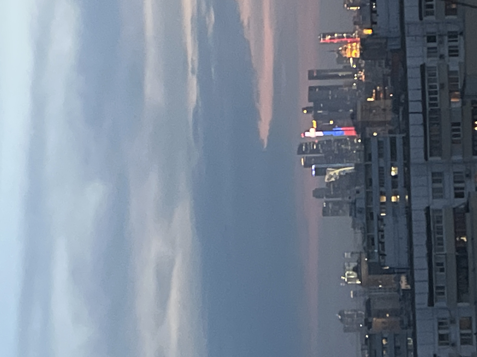 A distant cityscape is captured under a twilight sky. The sky is filled with a gradient of blues and grays, with subtle hints of pink near the horizon. The city skyline is dominated by modern skyscrapers, some of which are illuminated with colorful lights, including one building displaying the Russian flag colors. In the foreground, there are rows of apartment buildings with visible windows, some of which are lit, suggesting habitation. The image appears to be taken from an elevated position, looking out over the urban landscape. The overall atmosphere is one of dusk in a large metropolitan area.
