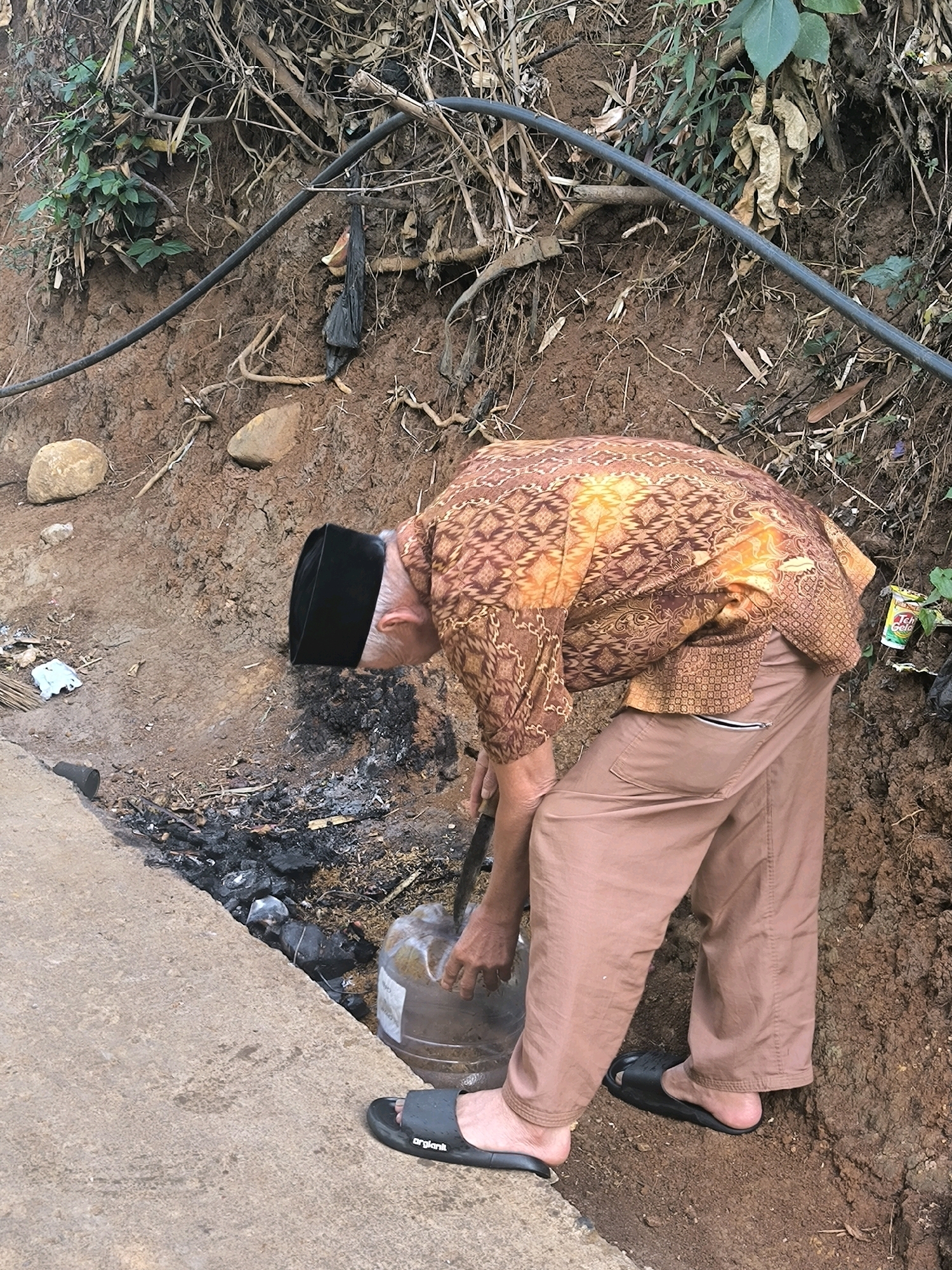 An older man wearing a black peci and a patterned brown and orange batik shirt is bent over, using a stick to agitate something in a large, clear plastic water jug. He is wearing light brown pants and black slide sandals. The jug is positioned near a patch of blackened earth, suggesting a fire or waste has been present. The scene appears to be outdoors, on a slope of reddish-brown soil with dry branches and green foliage. A black pipe runs diagonally across the upper portion of the image, secured by a wire. In the background, a can of "Teh Gelas" is visible. The ground is uneven, with loose soil and some rocks. A concrete edge is visible on the left side of the image. The overall impression is a rural or semi-rural setting in Cilengkrang, Indonesia.