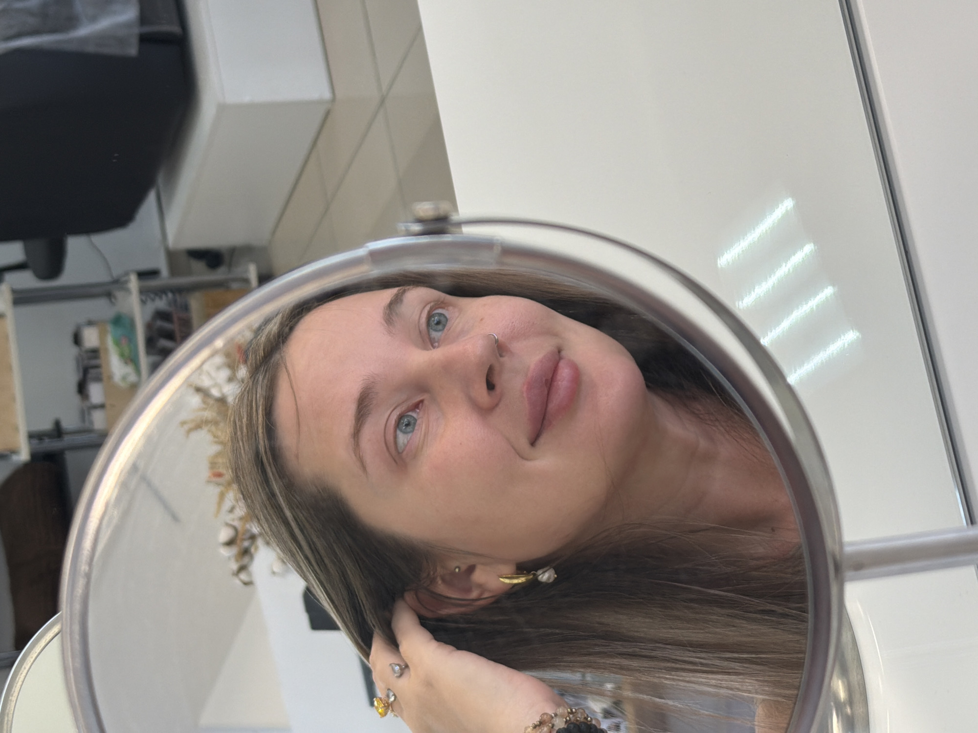 A woman is reflected in a circular makeup mirror. She has fair skin, light brown hair with blonde highlights, and blue eyes. She is wearing a nose ring and earrings. Her hand is near her ear, possibly adjusting her hair. Rings are visible on her fingers, and a bracelet adorns her wrist. The background is nondescript, with hints of a salon or dressing room environment including a white wall, possibly in Chaikovsky, Russia.