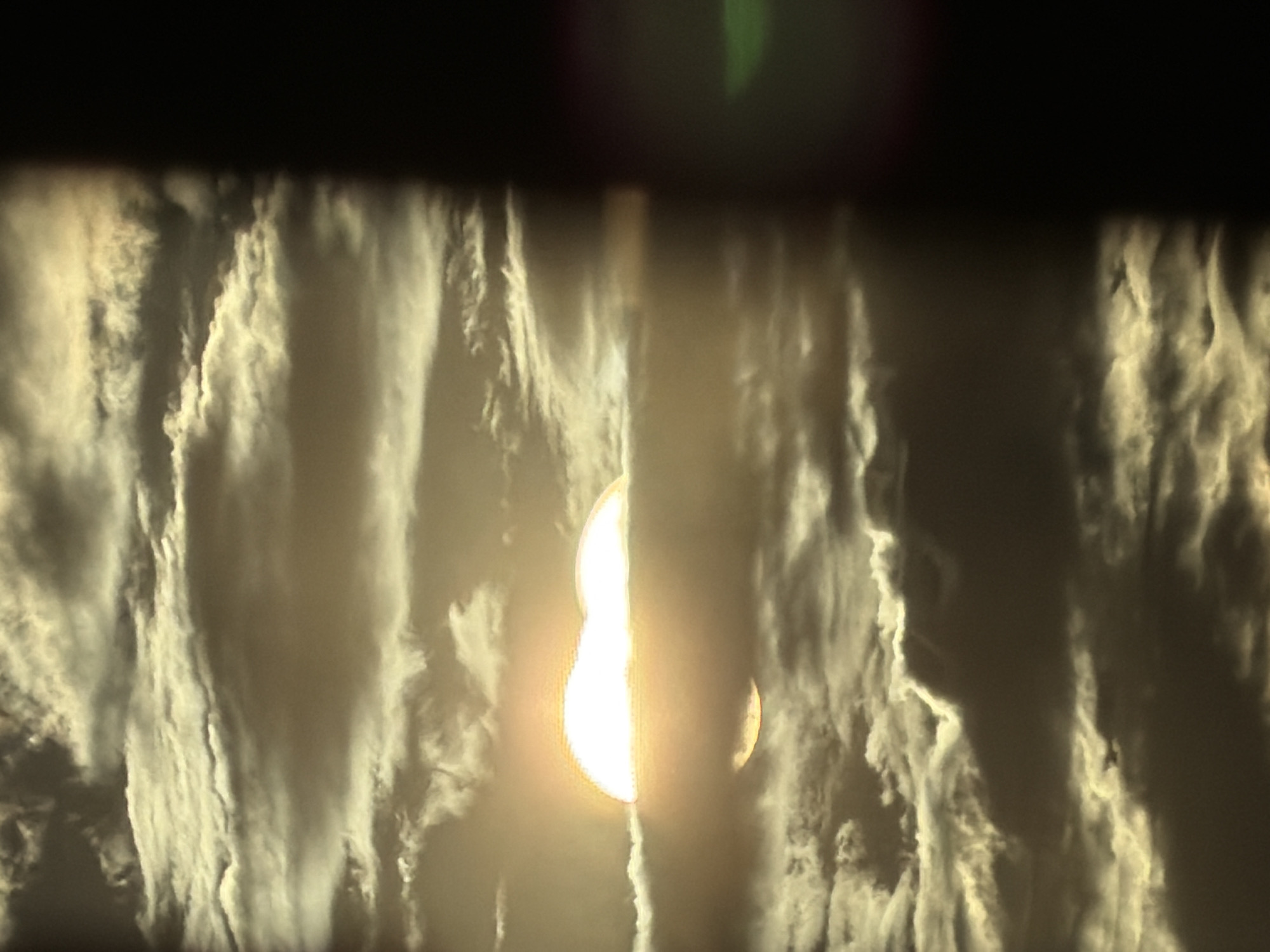 The image depicts a dramatic view of the sun partially obscured by thick, textured clouds. The sun appears as a bright, luminous orb, with a significant portion of it covered by a dark, cloud-like formation that creates a stark contrast. The clouds themselves are voluminous and wispy, with visible striations that suggest movement and depth. They are illuminated from behind by the sun, creating a radiant glow along their edges and highlighting their intricate textures.
The scene evokes a sense of awe and perhaps a touch of mystery, with the sun's light struggling to break through the dense atmospheric conditions. The overall color palette is dominated by warm tones of yellow and white from the sun and its light, contrasted with darker grays and blacks in the shadowed parts of the clouds. The composition is abstract, focusing on the interplay of light and shadow within the cloudscape. The upper portion of the image is darker, suggesting either a late hour or a deep shadow cast by an unseen element. There are no discernible people, objects, or explicit text within the frame. The overall impression is that of a powerful natural phenomenon, likely a sunrise or sunset witnessed through challenging weather conditions.
