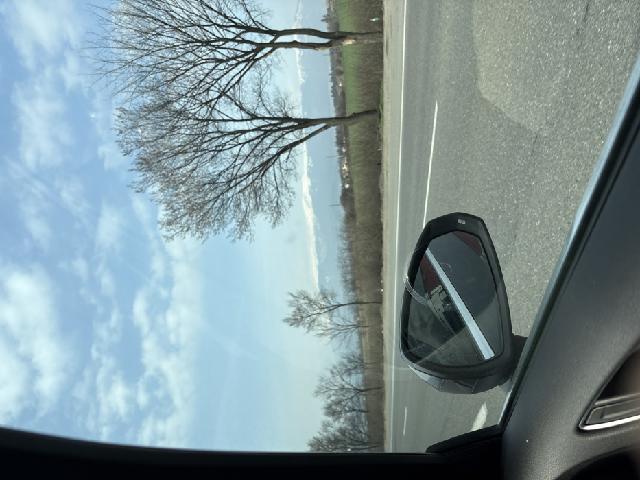 The image depicts a landscape captured from the interior of a vehicle in the city of Kazichene, Bulgaria. The primary view is through the vehicle's passenger-side window, revealing an exterior scene.
In the foreground, the dark gray or black interior door panel and window frame of the vehicle are visible on the left and bottom edges, with a metallic door handle partially present in the lower-left corner. A prominent black exterior side mirror is positioned in the lower-center, reflecting a white object with a distinct red horizontal stripe, indicative of another vehicle or a passing structure, alongside segments of the sky and the road.
The external environment features a paved asphalt road running horizontally across the mid-ground, marked with a solid white line delineating the shoulder. Beyond the road, the terrain consists of brown, possibly barren, fields interspersed with green patches. Numerous leafless deciduous trees with dark, intricate branch structures line the road and extend into the background. In the mid-distance, a cluster of small buildings is visible. The background is dominated by a range of snow-capped mountains under a light blue sky dotted with scattered white cumulus clouds. Faint linear contrails or streaks are discernible in the upper sky. No individuals or explicit interactions are visible.