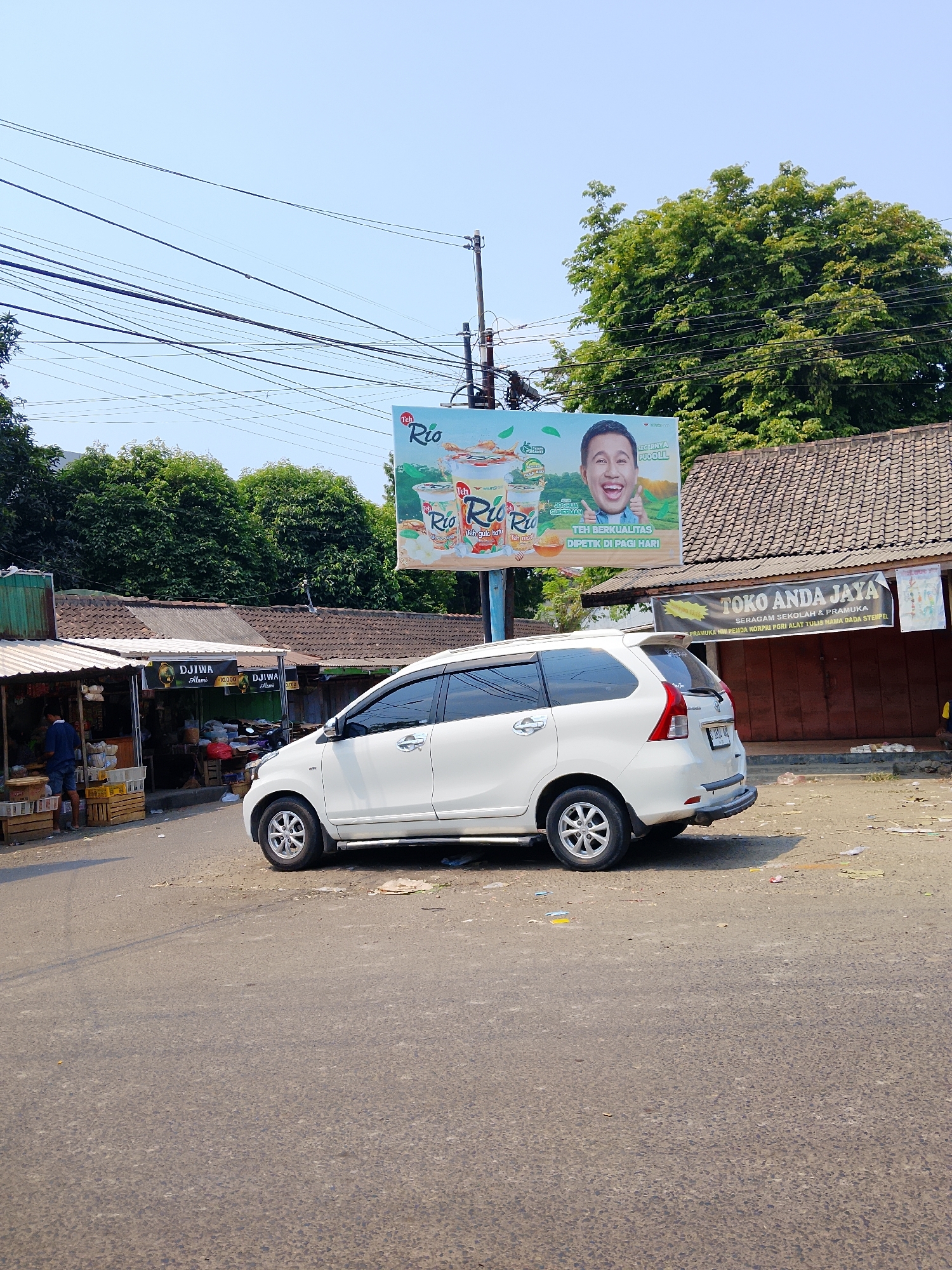 The image shows a street scene in Ngelo, Indonesia, on a sunny day. A white minivan is parked on the side of the road, facing away from the camera. In the background, there are several small shops with tiled roofs. A large billboard advertising "Teh Rio" is visible above the shops. There are also power lines and trees in the background. A person is standing near one of the shops, and another person is visible inside a shop. The ground is paved with asphalt and appears to be a bit dusty.