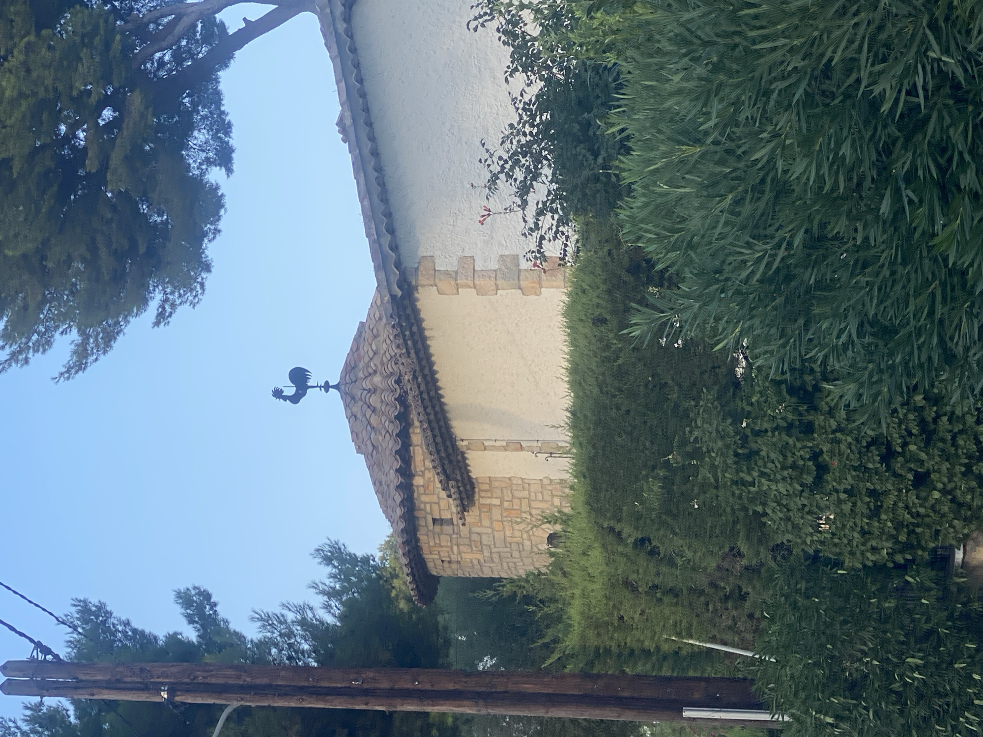 The image shows a building with a tiled roof, adorned with a weathervane shaped like a rooster. The building appears to be made of white stucco with stone accents. It is surrounded by lush green vegetation, including hedges and trees. In the foreground, a wooden utility pole with wires is visible on the left side of the frame. The sky above is a clear blue. The overall scene suggests a residential or possibly a small commercial property in a well-maintained, possibly Mediterranean, setting. Without further context, the specific location in Antibes, France, cannot be determined from the image alone.