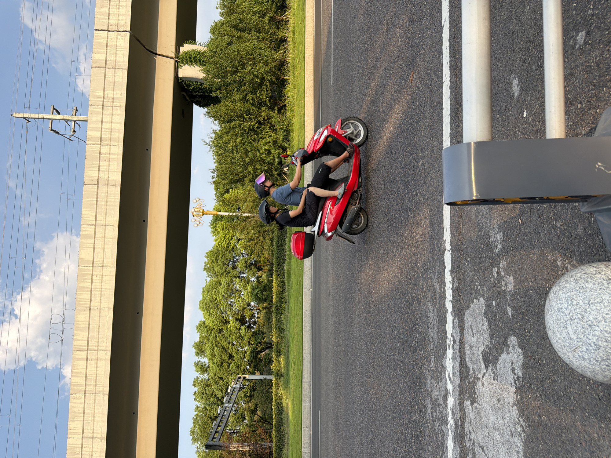 Two individuals, a driver and a passenger, are riding a red electric scooter along a paved road under an elevated concrete structure. The driver, wearing a blue shirt and dark shorts, operates the scooter, while the passenger, in a black t-shirt and black shorts, sits behind them. Both individuals are wearing helmets for safety. The scooter has a red storage box mounted on the rear. The road is bordered by a grassy median and lush green trees. Overhead, power lines and poles are visible against a sky with scattered clouds. The scene appears to be in a suburban or semi-urban area, possibly in Jiangjin, China, given the context. The lighting suggests it is daytime, with sunlight casting shadows. In the foreground, part of a guardrail and a large rounded stone are visible.
