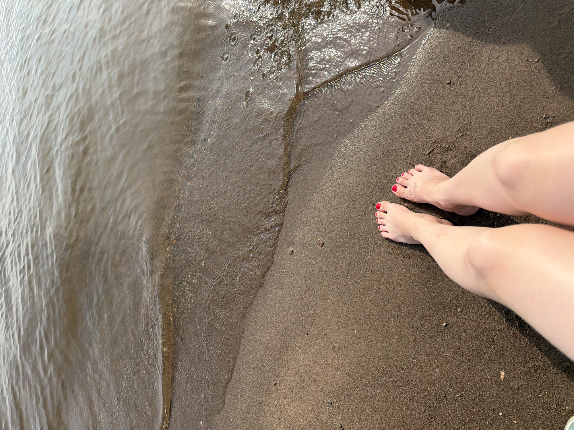 The image shows a pair of bare feet with red nail polish standing on wet sand at the edge of a body of water. The water is calm with small ripples. The sand appears to be dark brown and slightly wet. The overall lighting suggests it is daytime. The location is the Neva River beach in the city of Krasny Bor, Russia.