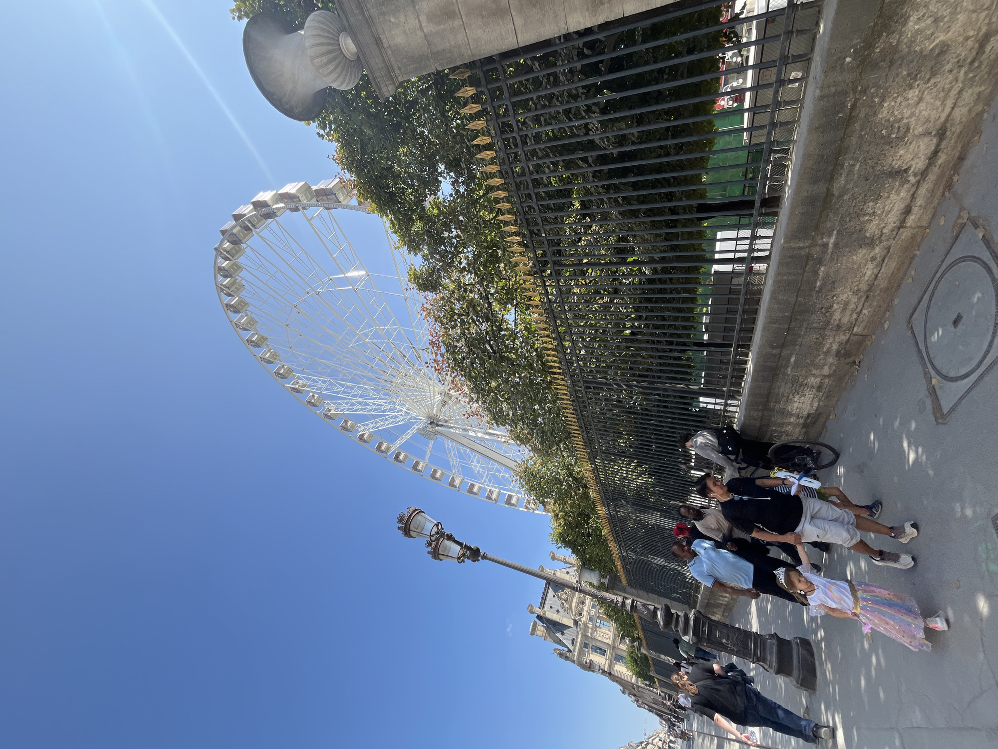 A white Ferris wheel is a prominent feature in this image, set against a clear blue sky on a sunny day. The Ferris wheel, with its many white gondolas, is a large structure that dominates the upper left portion of the frame. In the foreground, a group of people are walking along a paved sidewalk. A black metal fence runs along the right side of the image, with trees and foliage visible behind it.
The people in the foreground appear to be tourists. They are dressed in casual summer clothing. One person is pushing a stroller. The group includes adults and children. The mood seems relaxed and leisurely, indicative of a sightseeing excursion. A lamppost with multiple lights stands near the sidewalk. In the background, a building with architectural details is visible. The overall setting suggests an urban environment, likely a park or a public area in a city.
The time of day appears to be midday, given the bright sunlight and the shadows cast on the ground. The weather is fair and sunny. Although the location is identified as Paris, France, no specific landmarks are clearly identifiable beyond the Ferris wheel, which could be the Grande Roue de Paris. There is no visible text in the image.
