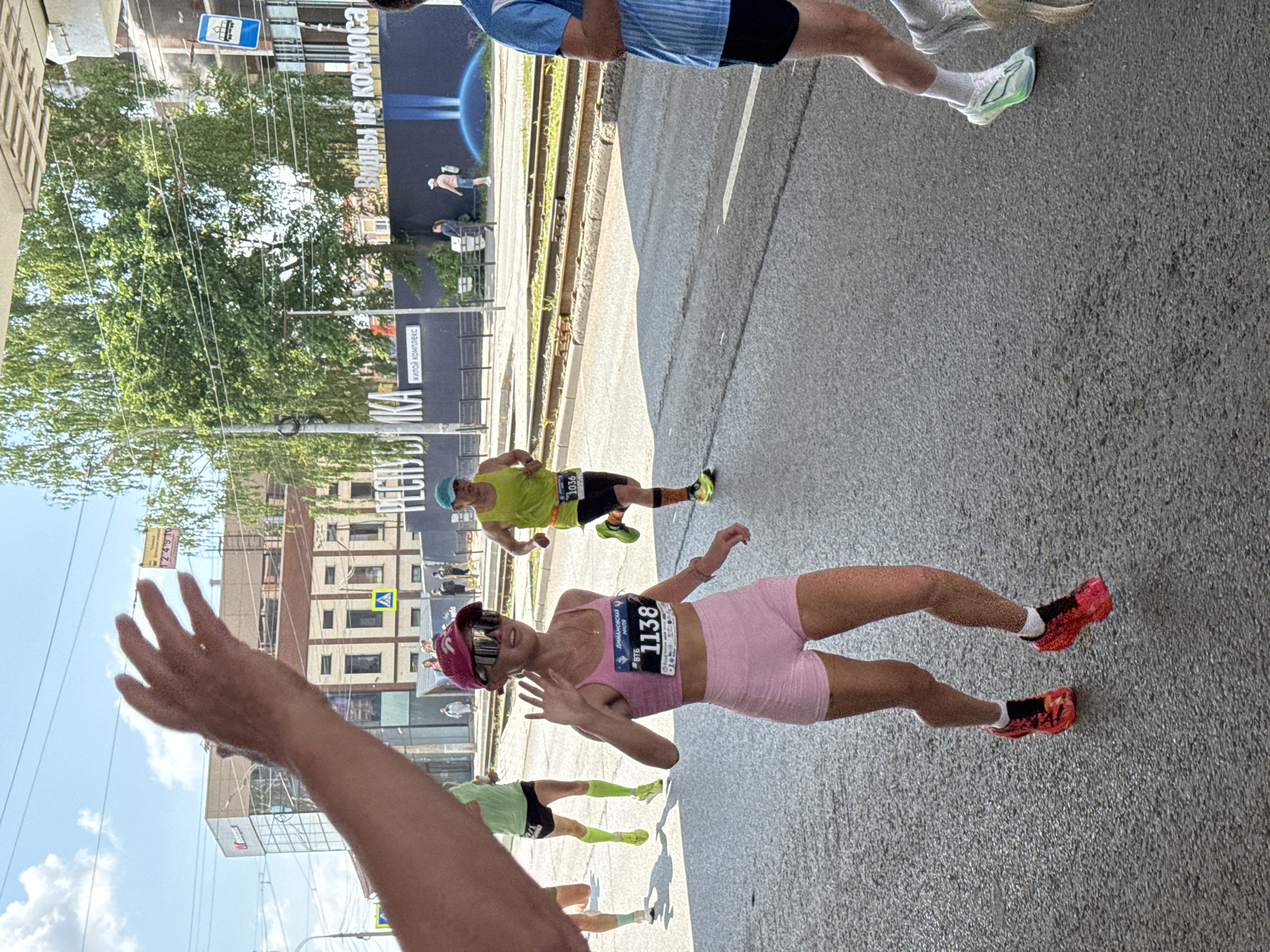 In Izhevsk, Russia, a group of runners participate in a marathon on a sunny day. The foreground features a female runner in a pink sports bra and matching shorts, with a bib number 1138. She is smiling and waving at the camera, her right hand held up. Behind her, other runners are visible, including a male runner in a neon green tank top with bib number 1036. The background shows city buildings, trees, and tram tracks. A large banner with Russian text is visible on a building, one part reading "ВИДНЫ ИЗ КОСМОСА" (Visible from space). The ground is paved asphalt, characteristic of a city street.