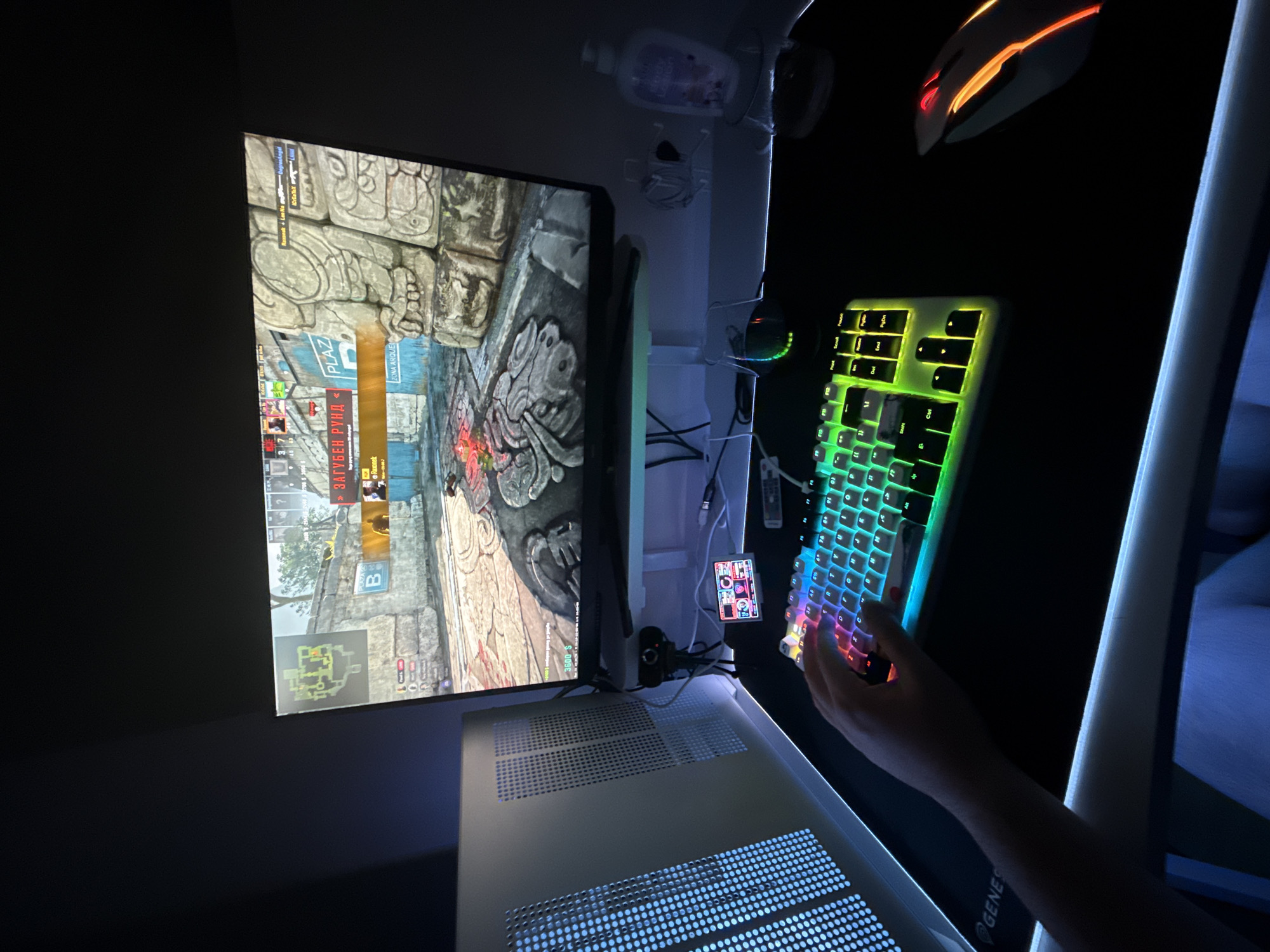 A low-angle, dimly lit image shows a person playing a video game on a computer. The game displayed on the monitor appears to be a first-person shooter, featuring a map titled "Zona Arque". Various in-game elements are visible, including the player's health, money ($3600), and the objective "B". In the HUD, it appears the language is Bulgarian, and the message reads "ЗАГУБЕН РУНД" which translates as "LOST ROUND".
The person's hand is visible as they interact with a brightly lit, multi-colored keyboard. An orange and white gaming mouse sits to the right of the keyboard, suggesting active gameplay. The environment appears to be a personal computer setup, with a computer case visible beneath the monitor, featuring illuminated ventilation holes. Cables and other accessories are also visible around the monitor and desk. The room is generally dark, with the illuminated keyboard and monitor providing the primary sources of light.