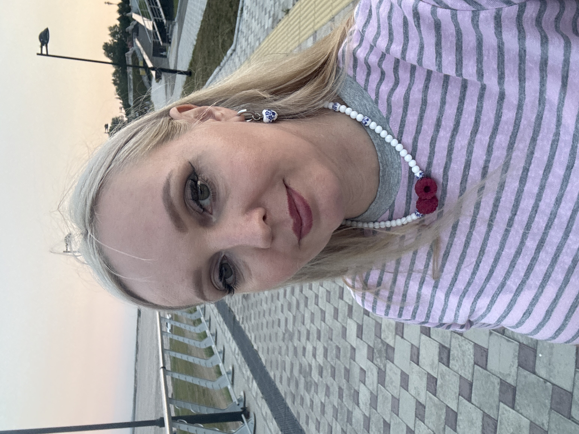 A medium shot selfie shows a fair-skinned Caucasian woman with shoulder-length blonde hair, wearing a pink and gray striped long-sleeved shirt and a white beaded necklace with blue accents and two red raspberry-like charms. She has on eye makeup and lip color, and she is wearing a hoop earring with a blue and white patterned heart charm. She is standing on a paved walkway with a checkered pattern of light and dark gray bricks. To her left, a metal railing lines a pathway or embankment. In the background, a body of water is visible, and beyond that, trees and a streetlamp. The sky is a pale pink and orange, suggesting it is either sunrise or sunset. The environment appears to be an urban park or promenade in Komsomolsk-on-Amur, Russia, given the specific architectural style of the railing and paving that is characteristic of the city's waterfront revitalization projects.