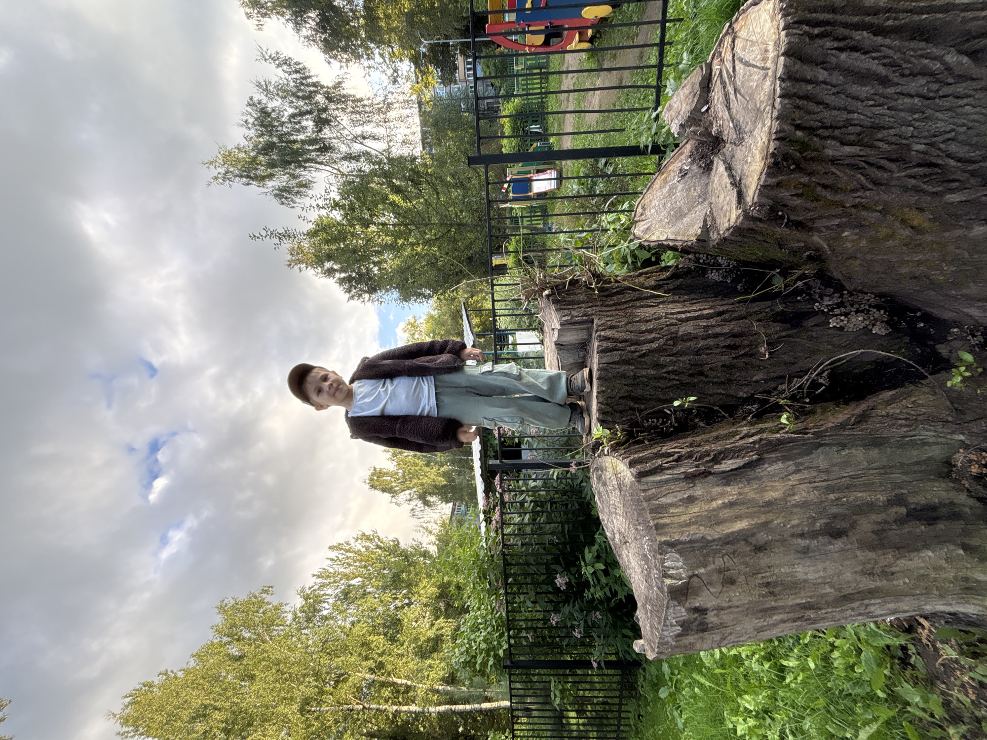 A young boy stands on a large tree stump in a park. He is wearing a light blue t-shirt, a brown zip-up jacket, and light green cargo pants, and a brown baseball cap. The tree stump is in the foreground, with a black metal fence and a playground visible in the background. Trees and buildings can be seen further in the distance, and the sky is overcast with clouds. The location is identified as Rusanovo, Russia.