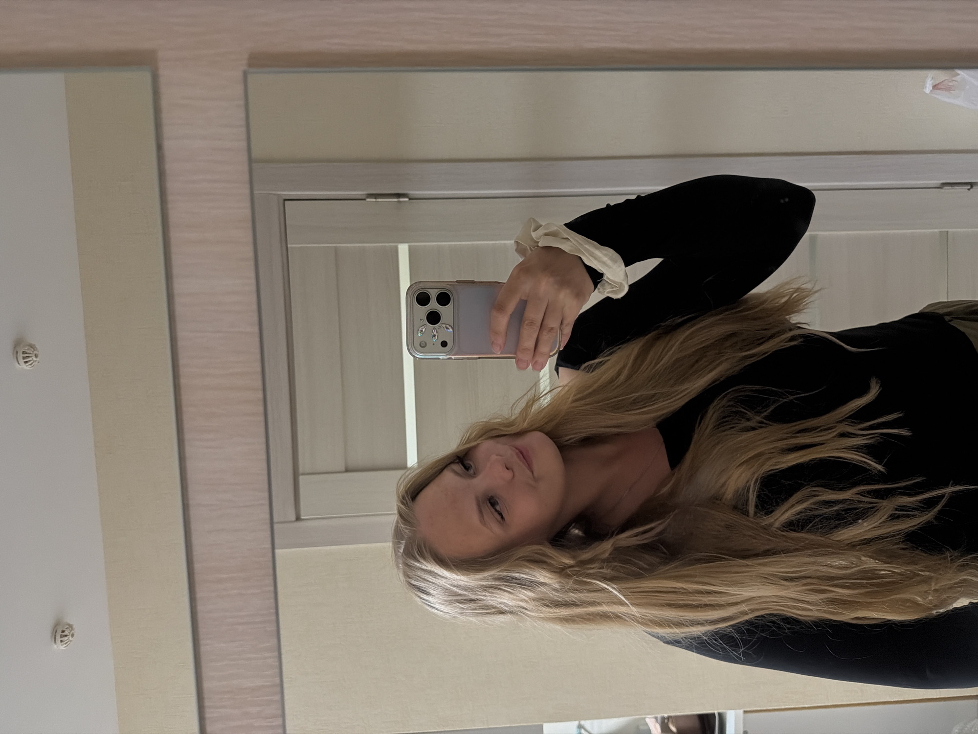 A young woman with long blonde hair is taking a mirror selfie in what appears to be a residential interior, possibly an apartment hallway or room in Izhevsk, Russia. She is wearing a black long-sleeved top with a white ruffled cuff. She is holding a smartphone in her right hand, angled to capture her reflection. The background shows a neutral-colored wall, a doorway with a light-colored door, and a ceiling with a smoke detector. The overall lighting suggests an indoor setting during the day.
