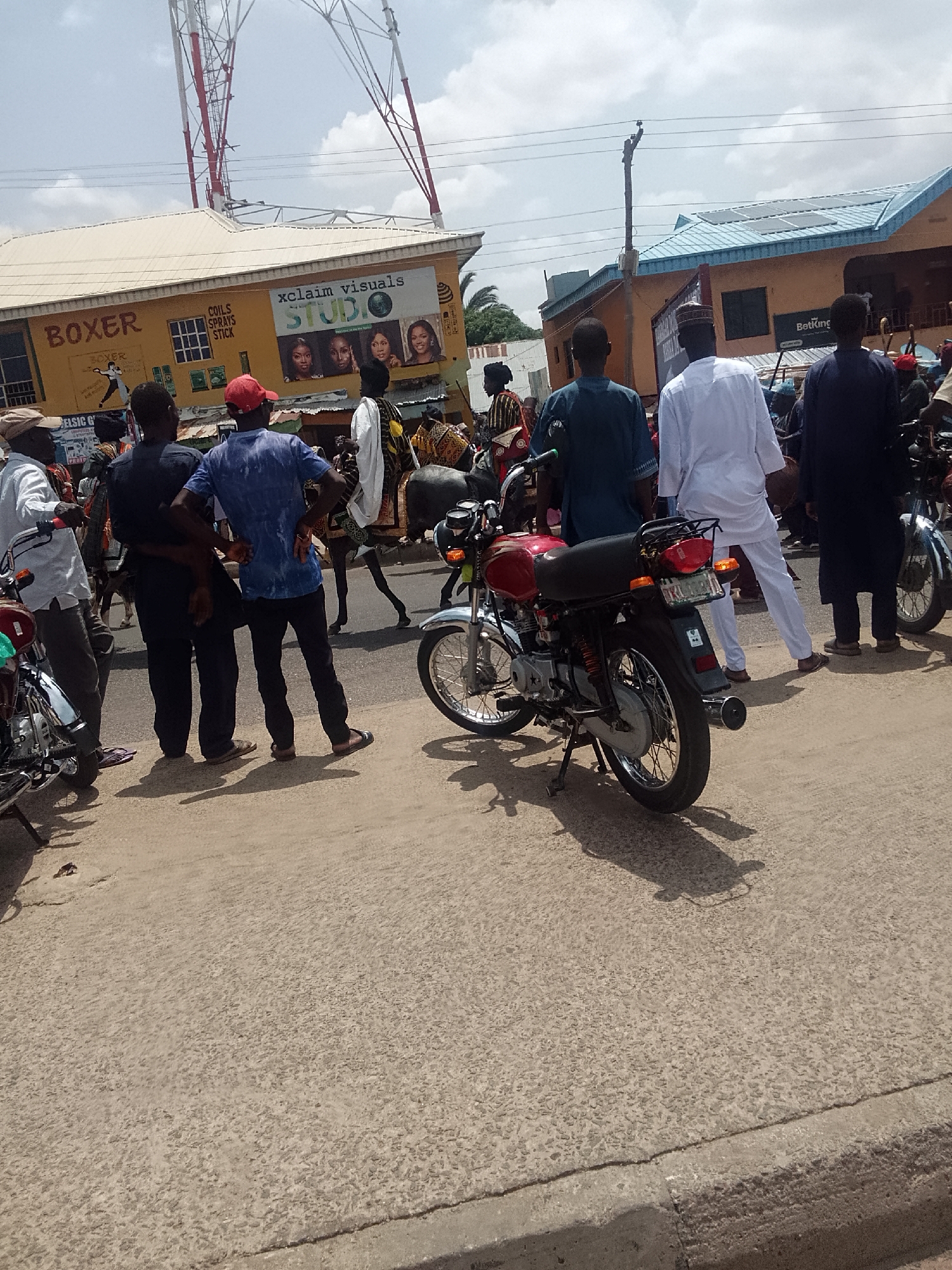 The image depicts a street scene in Arawa, Nigeria, with a group of people gathered on the side of the road. A red motorcycle is parked prominently in the foreground. A man riding a horse is walking across the street, surrounded by the crowd. Buildings line the street, including businesses with signs for "Boxer," "Xclaim Visuals," and "BetKing." A tall communication tower is visible in the background. The sky is partly cloudy.