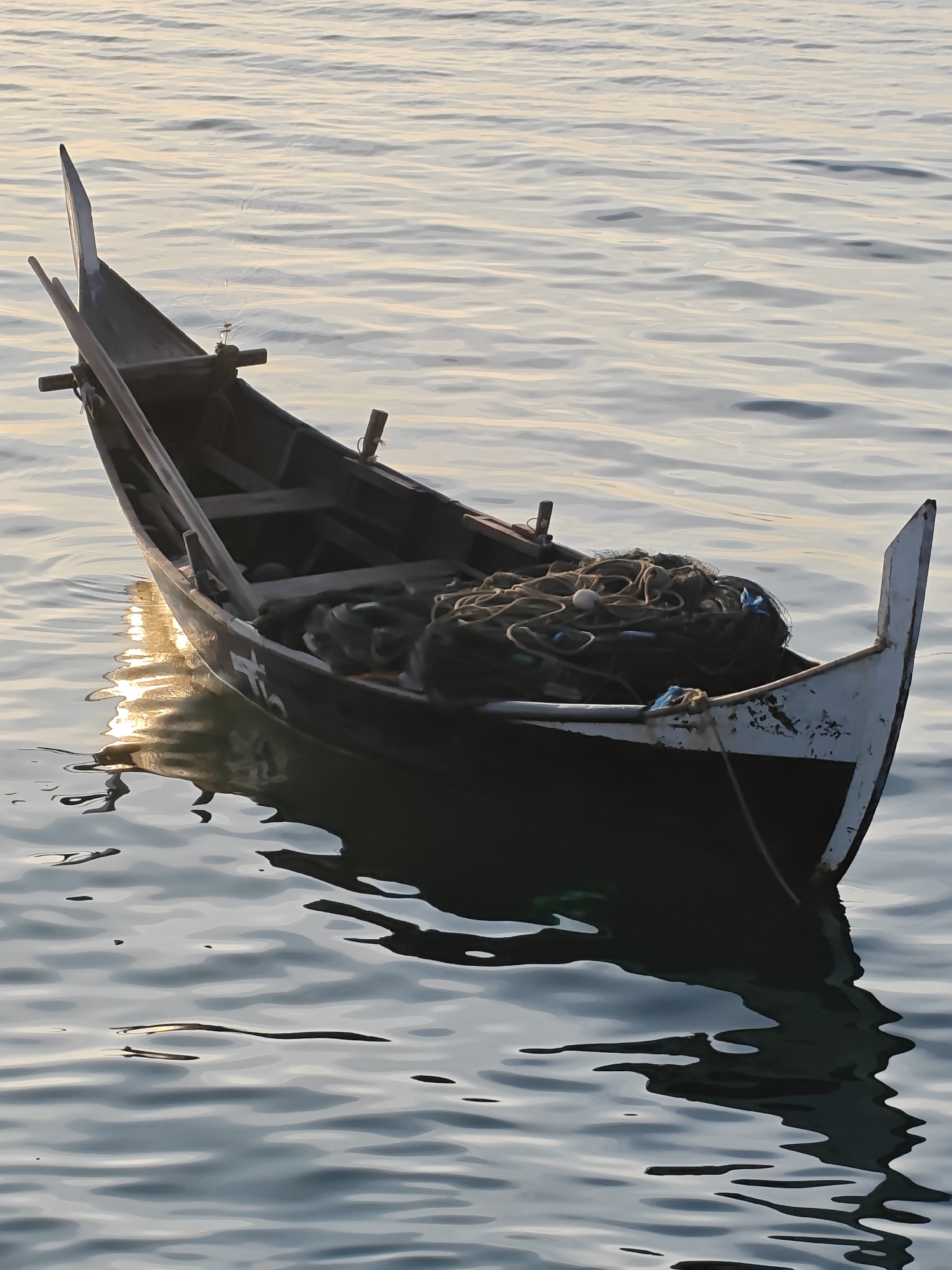 A wooden fishing boat is anchored in the water near the coast of Lhokseumawe, Indonesia. The boat is dark in color and filled with fishing nets. The water is calm with gentle ripples, reflecting the soft light of the setting sun. The scene captures a moment of tranquility and the livelihood of the local fishermen.