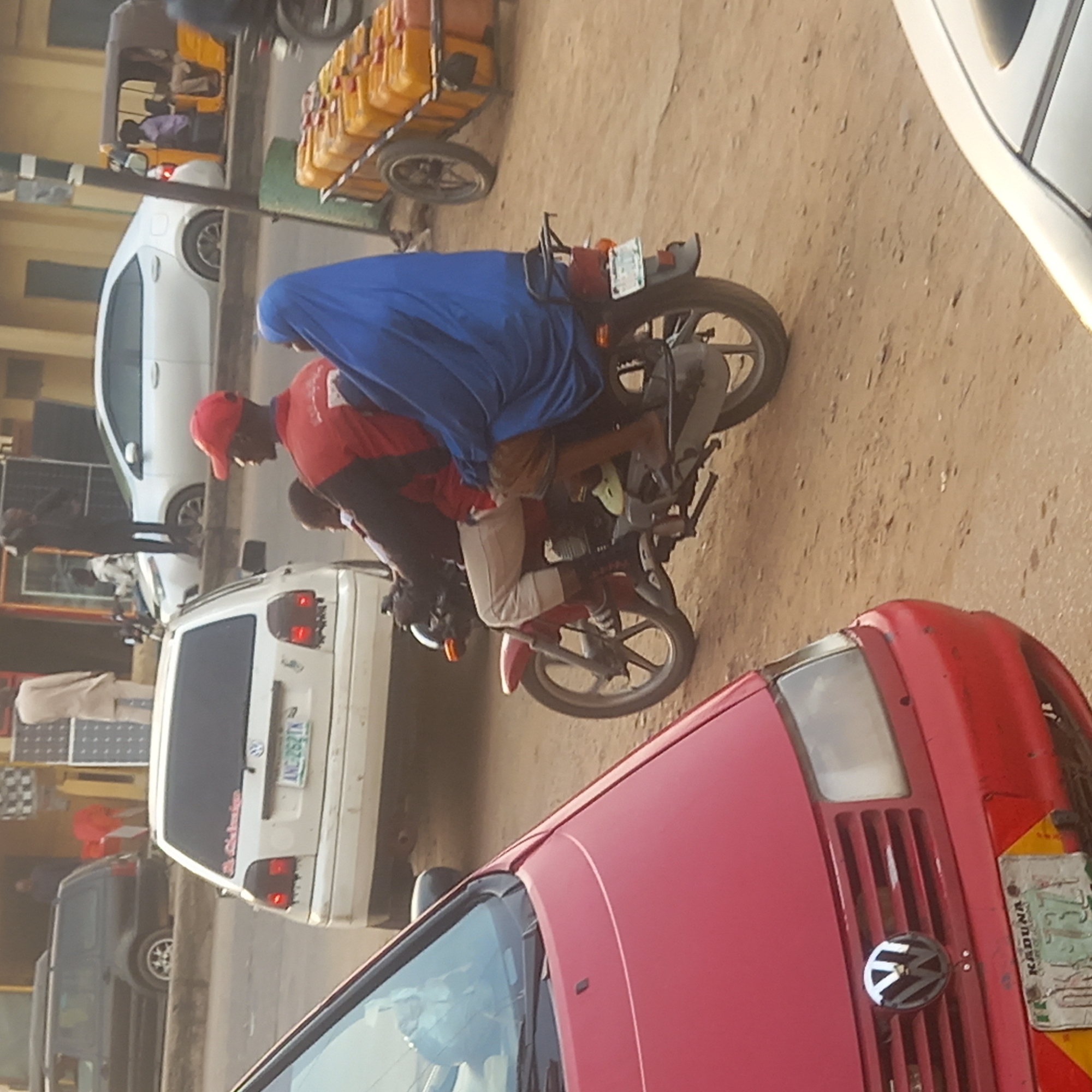 The image depicts a street scene in Dadi Riba, Nigeria. Two people are riding on a motorcycle. The driver is wearing a red shirt and a red cap. The passenger is covered in a blue cloth. A red Volkswagen car is in the foreground and a white van is behind the motorcycle. A yellow vehicle with canisters is further in the background.