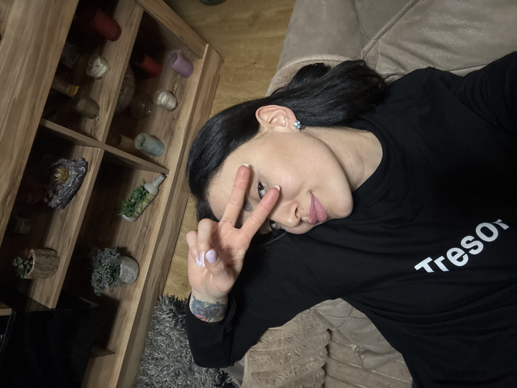 A person with black hair and a black shirt with the word "Tresor" on it is lying on a beige couch, holding up a peace sign. The person has an earring in their left ear, a tattoo on their left wrist, and painted light purple nails. To the left, there is a wooden shelf with various decorative items on it. The flooring is a light shade of brown and gray. The location cannot be determined from the information provided.