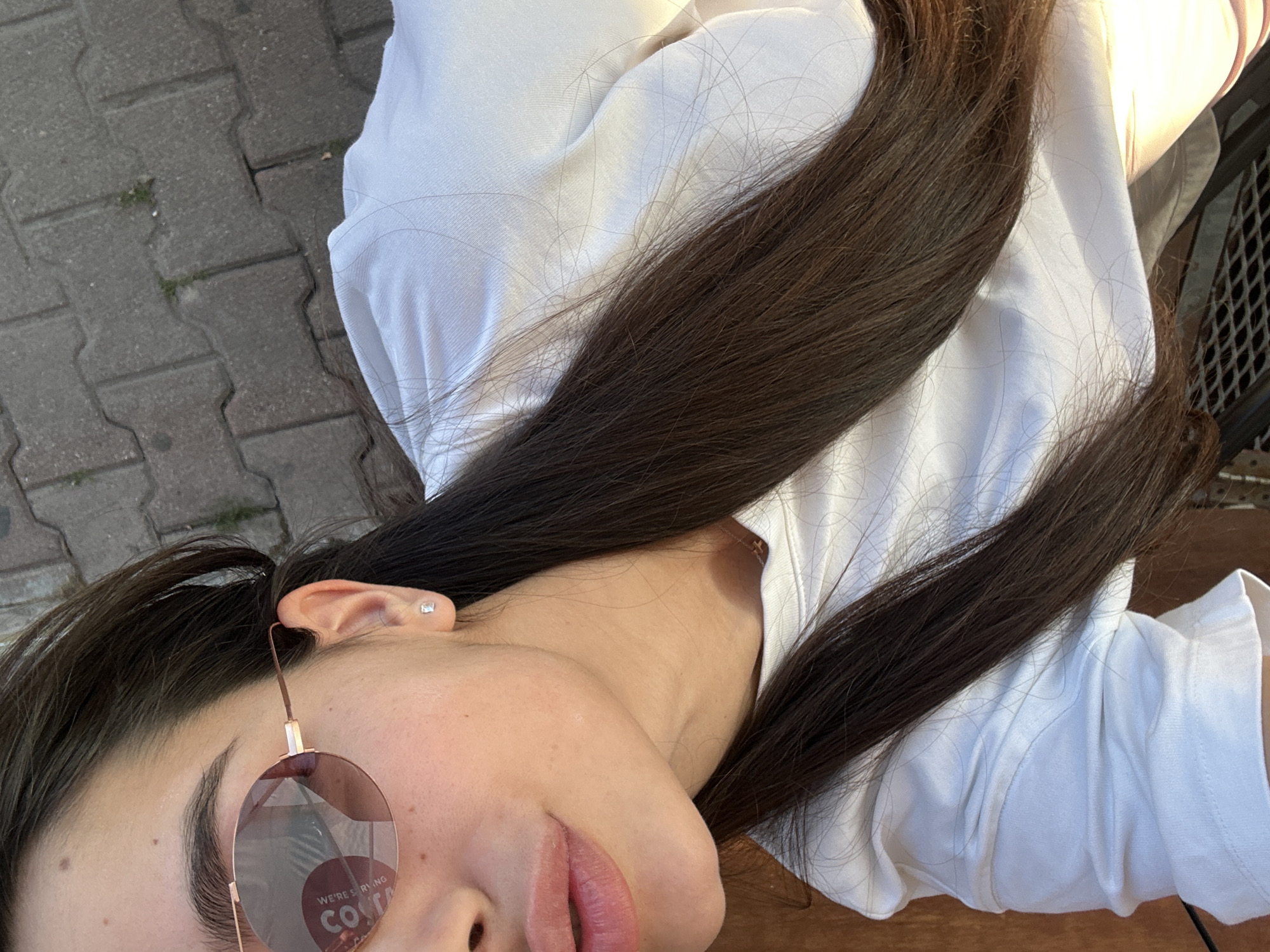 A close-up, high-angle shot shows a person, likely female, with dark brown hair, wearing a white shirt or top, and round sunglasses with a "We're serving Costa" design visible on the lens. A glimpse of a brick walkway appears on the left side. The person's left ear is visible, featuring a small earring. The location is presumed to be in the city of Plovdiv, Bulgaria.