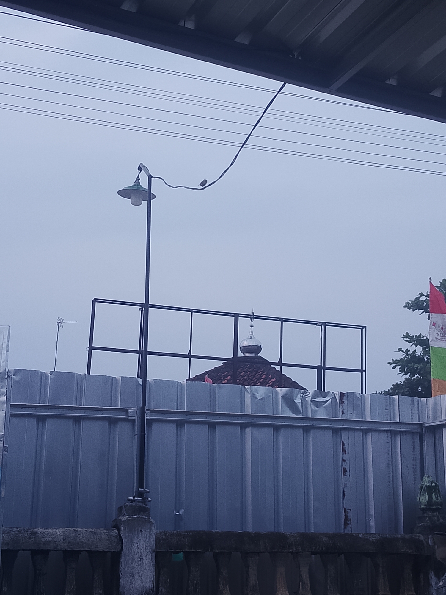 A photograph taken from a low vantage point, looking up towards a cloudy sky, depicts an outdoor scene with a temporary fence made of corrugated metal panels. A portion of a building with a tiled roof and a dome is visible above the fence. A black metal frame structure, possibly for a billboard or signage, is also present. A lamppost with a green lampshade stands on the left side of the image. A thin wire extends from the lamppost, with two small birds perched on it. The foreground shows a weathered stone railing or low wall with decorative elements, and a concrete post. The overall impression is of an urban or semi-urban environment, possibly undergoing construction or renovation, given the temporary fencing. The presence of a dome suggests a religious building, likely a mosque, given the location in Negeri Agung, Indonesia, known for its predominantly Muslim population. The flags visible on the right further suggest a location in Indonesia, where flags are commonly displayed. The weather appears overcast, contributing to a subdued light in the photograph.