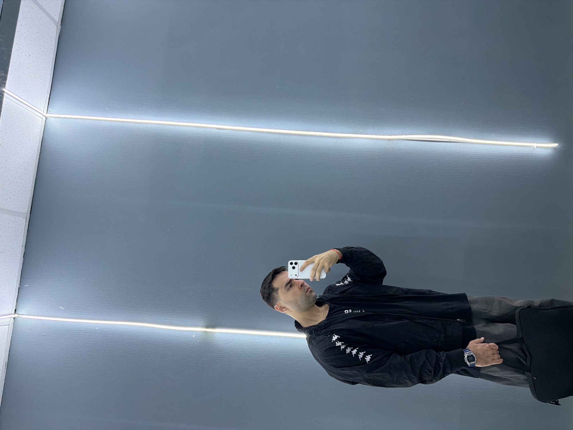 A man is holding a cell phone and taking a selfie in a space with a light gray ceiling. The ceiling features two linear LED light fixtures. The man is wearing a black Kappa jacket and a watch. The location is likely indoors, possibly in a shop or office. There is no information in the photo that indicates it is in Krasnoyarsk, Russia.