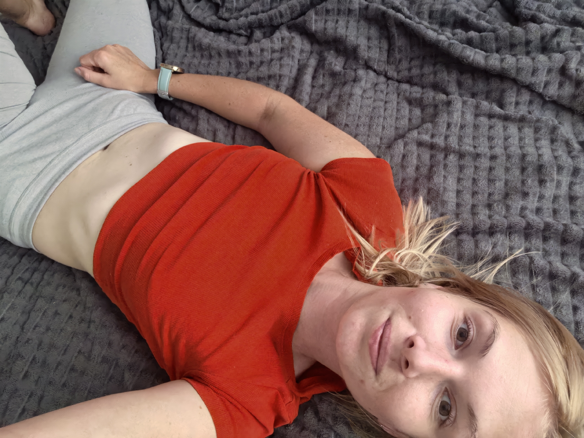 A woman is lying on a textured gray blanket, looking up at the camera. She is wearing a bright orange short-sleeved shirt and light gray leggings. A blue and gold smartwatch is visible on her left wrist. Her blonde hair is spread out around her head. She has a faint smile and her eyes are open, looking directly at the viewer. The overall lighting suggests it could be daytime, possibly indoors, as there are no strong shadows or indications of natural outdoor elements. The context provided indicates the location is Sterlitamak, Russia. No text is visible in the image.