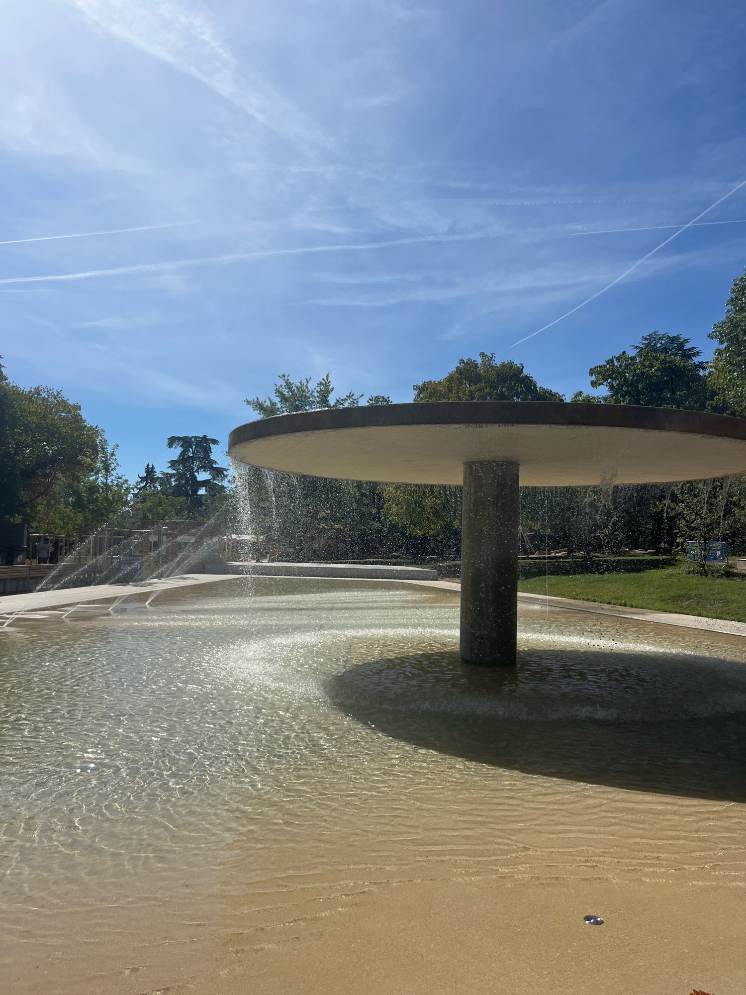 A modern fountain with a wide, flat canopy sits in the foreground, with water cascading over its edge. The water flows into a shallow pool with a sandy bottom. In the background, lush green trees line the perimeter of the fountain area. The sky above is a clear blue, with faint wisps of clouds and contrails from airplanes crisscrossing it. The overall scene suggests a sunny day in a park or public space, possibly a recreational area within the city of Geneva, Switzerland. There are no visible people in the immediate vicinity of the fountain, though some structures and benches are visible in the background, hinting at its use by the public.
