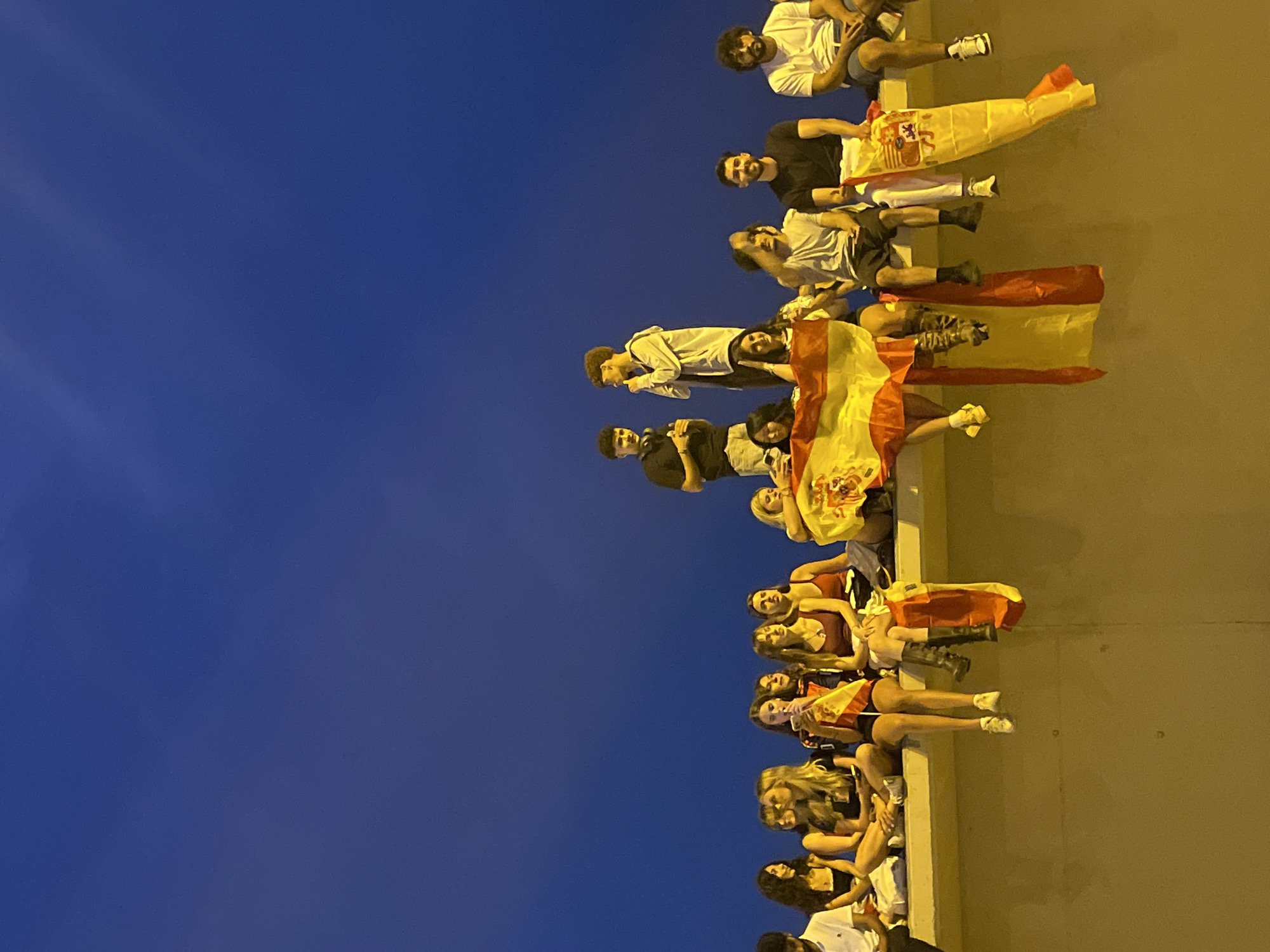 The image shows a group of young people sitting on a raised concrete ledge against a dark blue sky. Most of the individuals are facing forward and appear to be watching something. Several people are holding Spanish flags, some of which are draped over the ledge. The overall atmosphere seems casual and possibly celebratory. The perspective suggests the photo was taken from below and slightly to the side of the group. The lighting indicates it is either dusk or dawn. There are no discernible landmarks that specifically place the scene in Barcelona, Spain.
