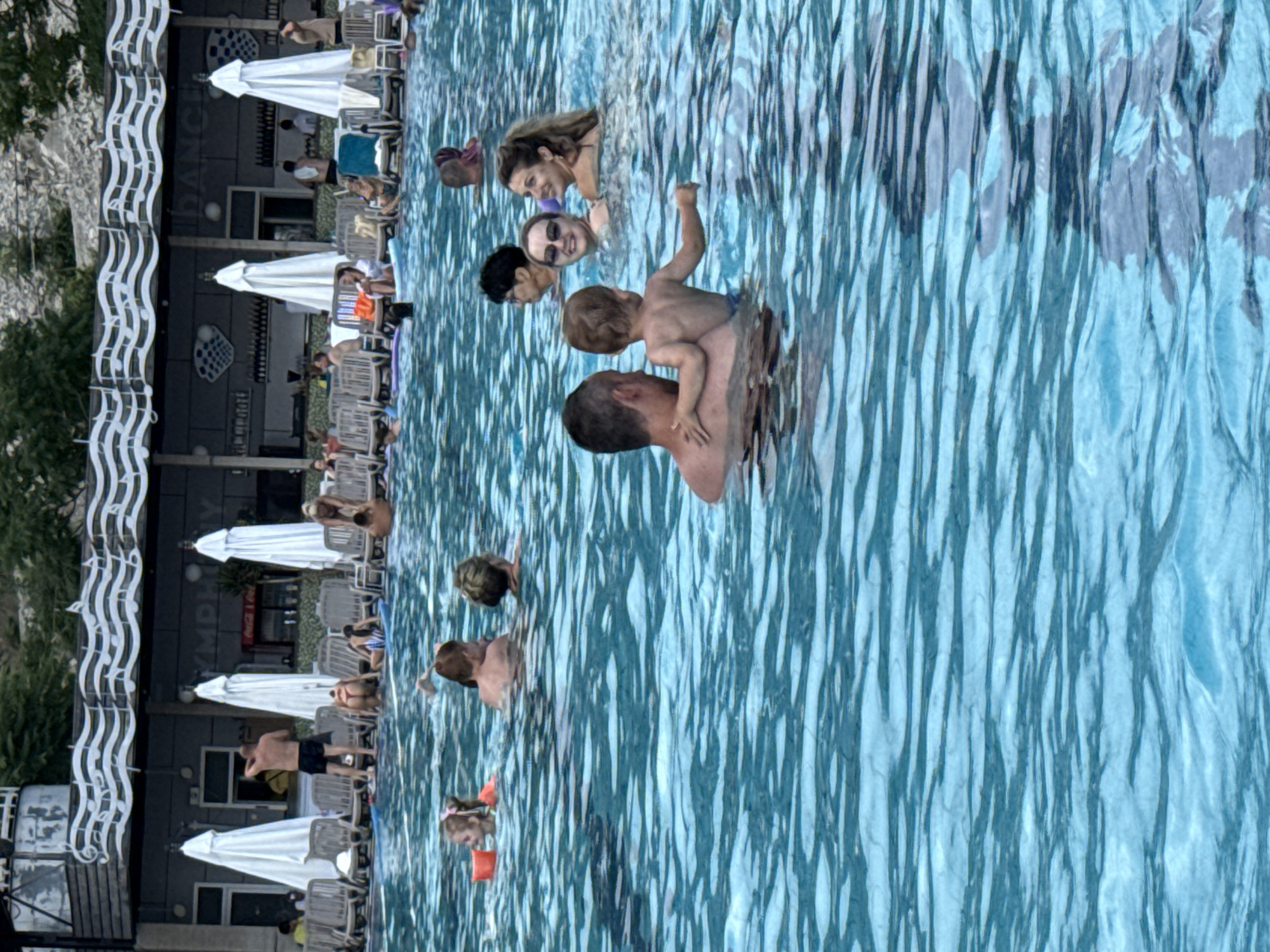 The image depicts a crowded outdoor swimming pool area. In the foreground, a man and a young child are in the water, with the child being held by the man. To their right, two women are in the pool, one wearing sunglasses. Further back, other people are swimming, lounging on chairs, and standing near a bar area. The background features a building with a decorative musical motif on its facade, along with umbrellas and lounge chairs arranged around the pool. The water is a vibrant blue, reflecting the surrounding environment. The scene appears to be a lively resort or hotel pool, possibly located in a sunny climate. The setting suggests a vacation or leisure activity, with many people enjoying the water and amenities.