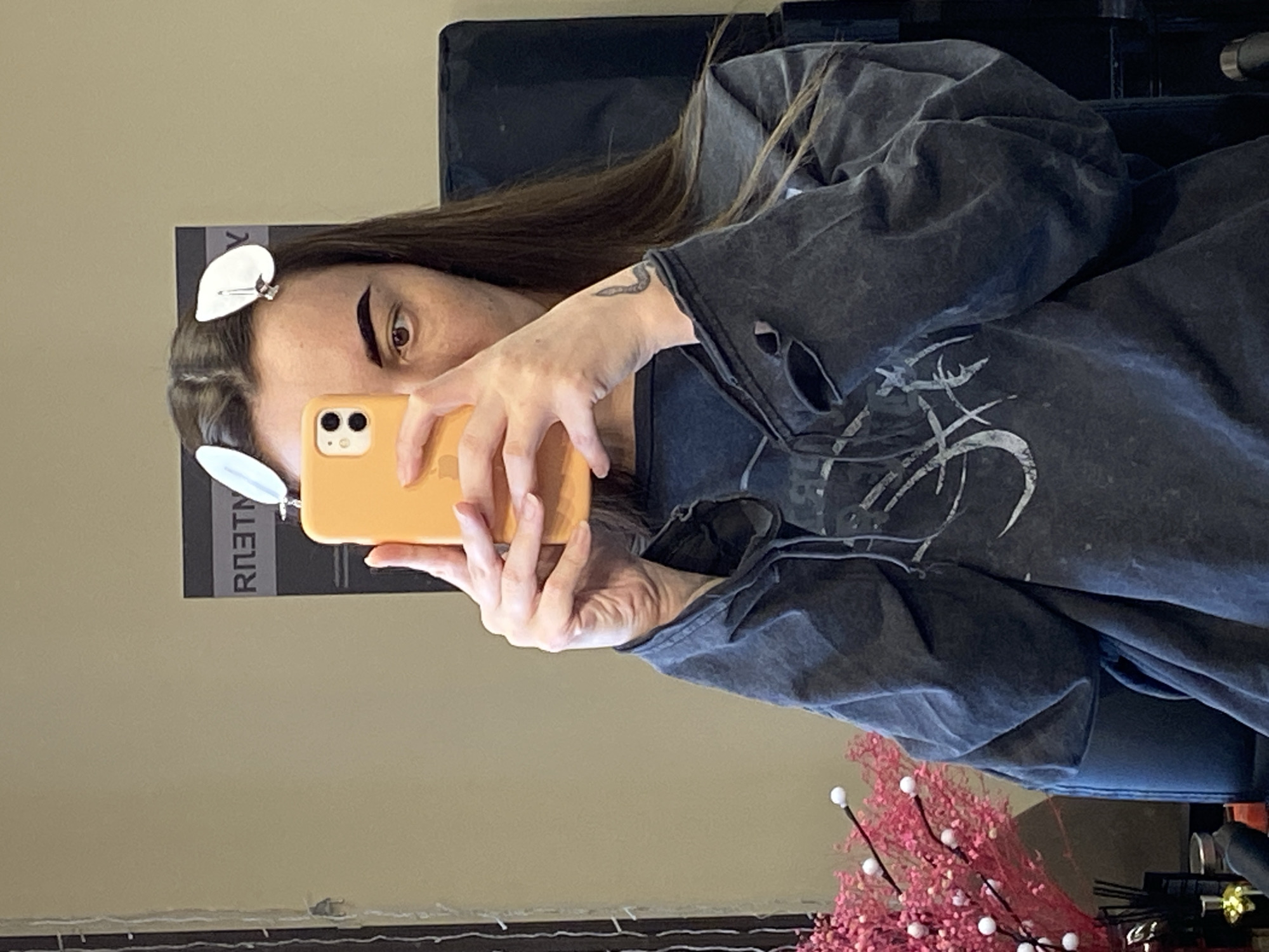 A woman with brown hair and pale skin is taking a selfie in a seated position. She has two white hair clips in her hair near her forehead. Her dark-colored shirt has a distressed design and holes in the sleeve. She's holding an orange smartphone in her hands, partially obscuring her face. A tattoo is visible on her left forearm. She is sitting in a chair in front of a light-beige wall. A small pink and white decorative floral arrangement and several objects are at the bottom right of the image. The location and city of Votkinsk, Russia, are not determinable from this image.