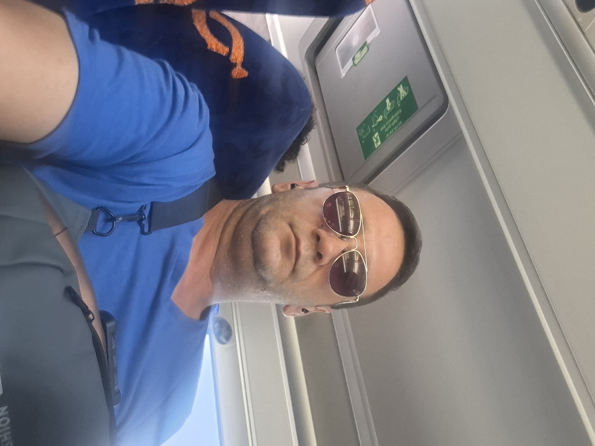 A man is taking a selfie while seated inside a vehicle, likely a bus or train. He is wearing aviator sunglasses and a blue t-shirt, with earbuds in his ears. The background shows the interior of the vehicle, including a ceiling panel with an "Emergency Exit" sign and a blue seat with an orange design. The man appears to be looking directly at the camera. The image was taken in Be'er-Sheva, Israel.