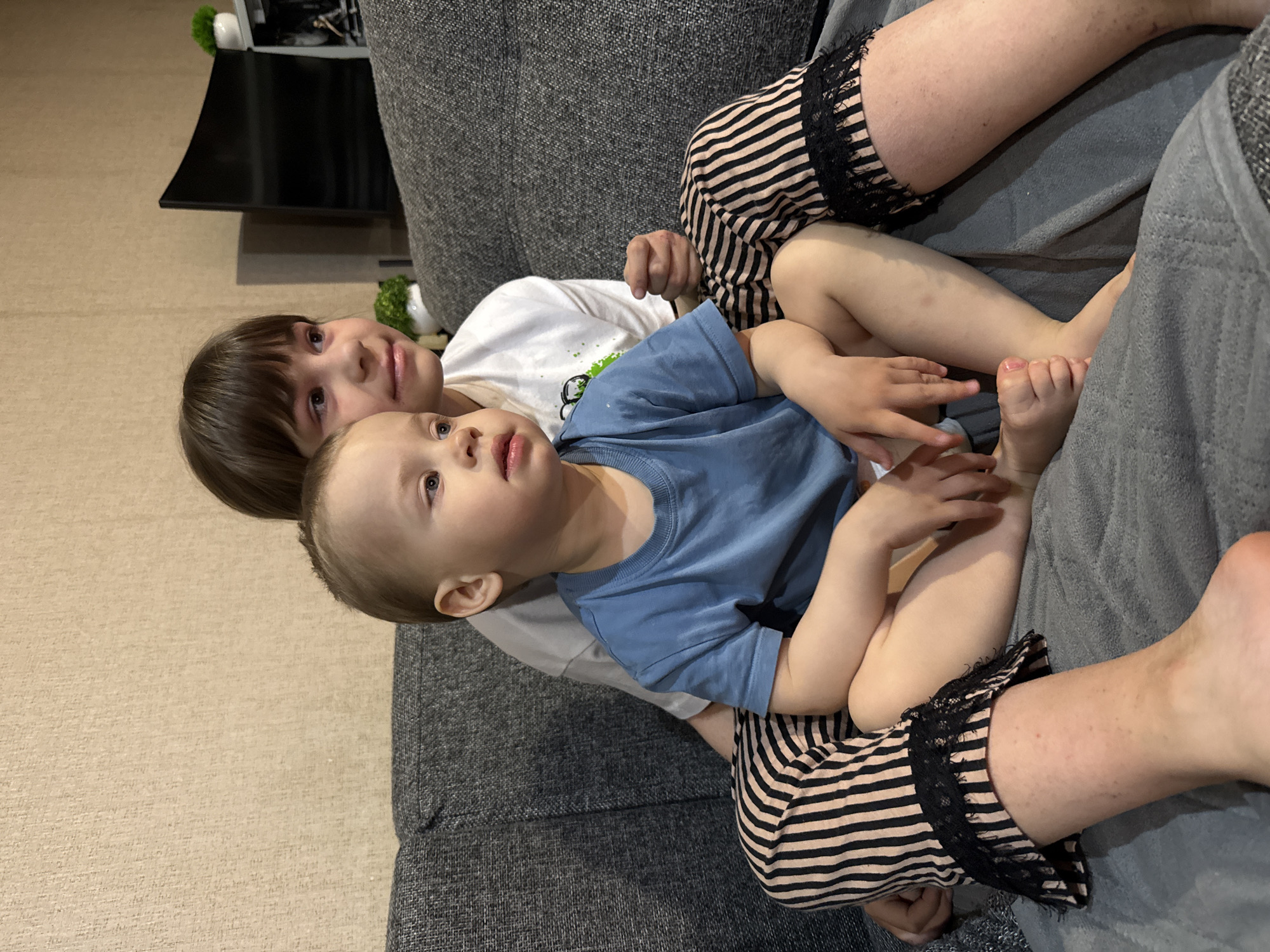 Two children are sitting on a gray upholstered sofa. One child, wearing a blue shirt, is in the foreground and looking upwards. Behind them, another child wearing a white shirt looks towards the camera. They are both situated on the lap of a person wearing striped shorts with a lace trim. The scene is indoors. Rusanovo, Russia is not discernible in the image.